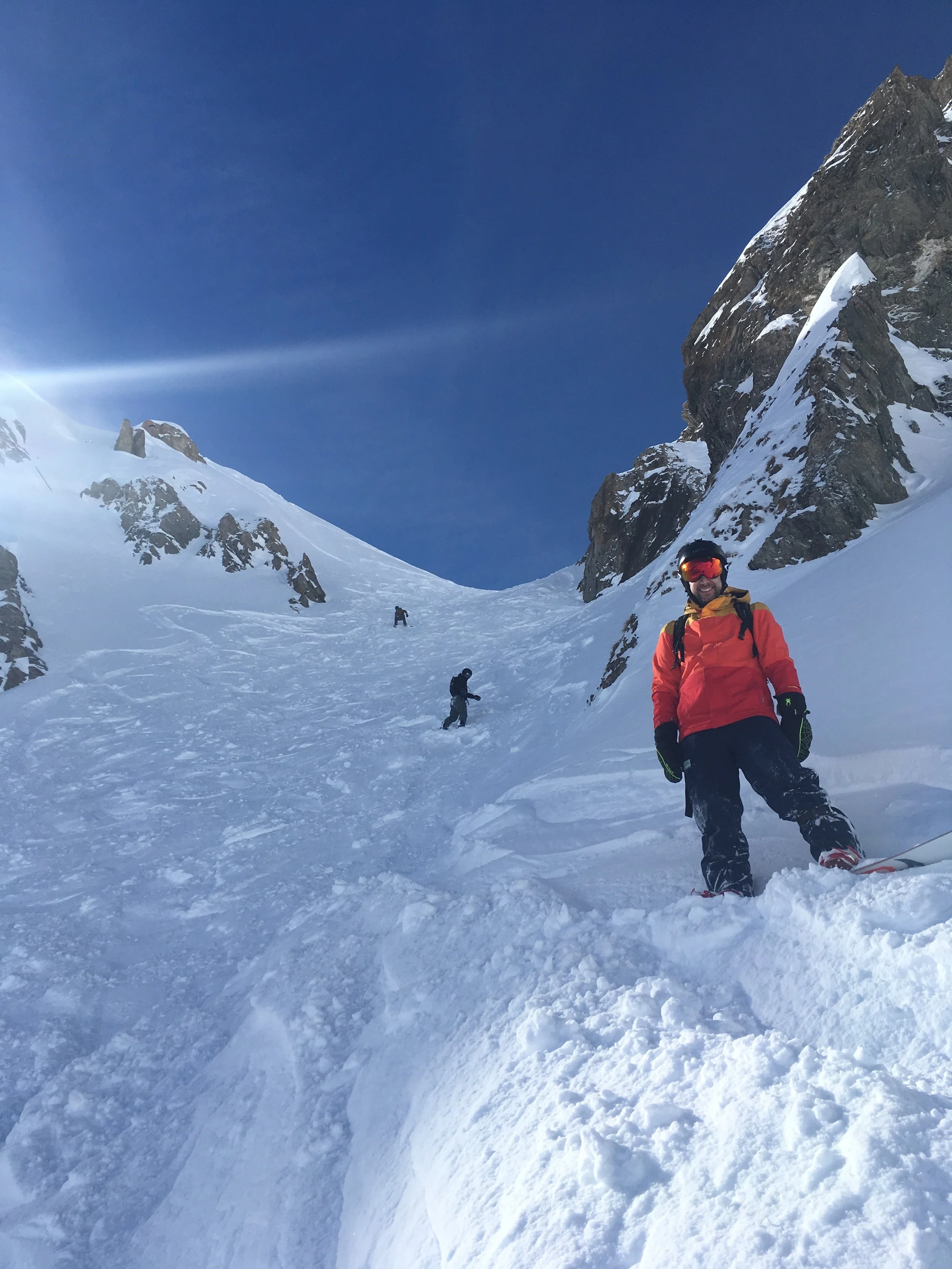 A group of people skiing or snowboarding on a snowy mountain slope with rocky cliffs and a clear blue sky.