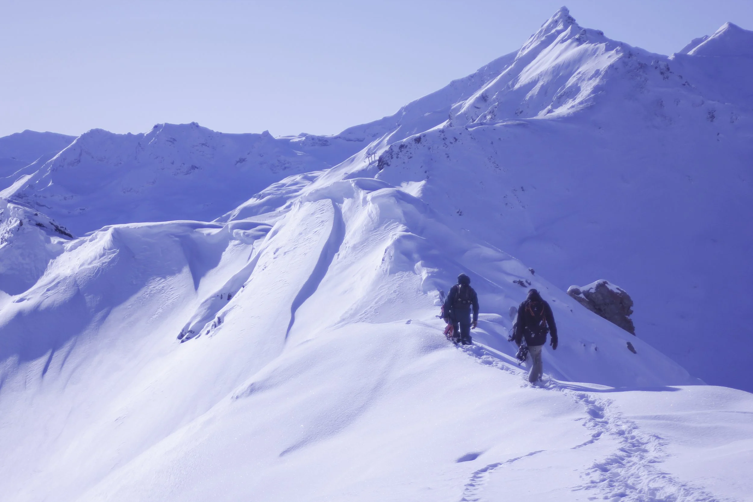 Three hikers walking through a snowy mountain landscape on a clear day.