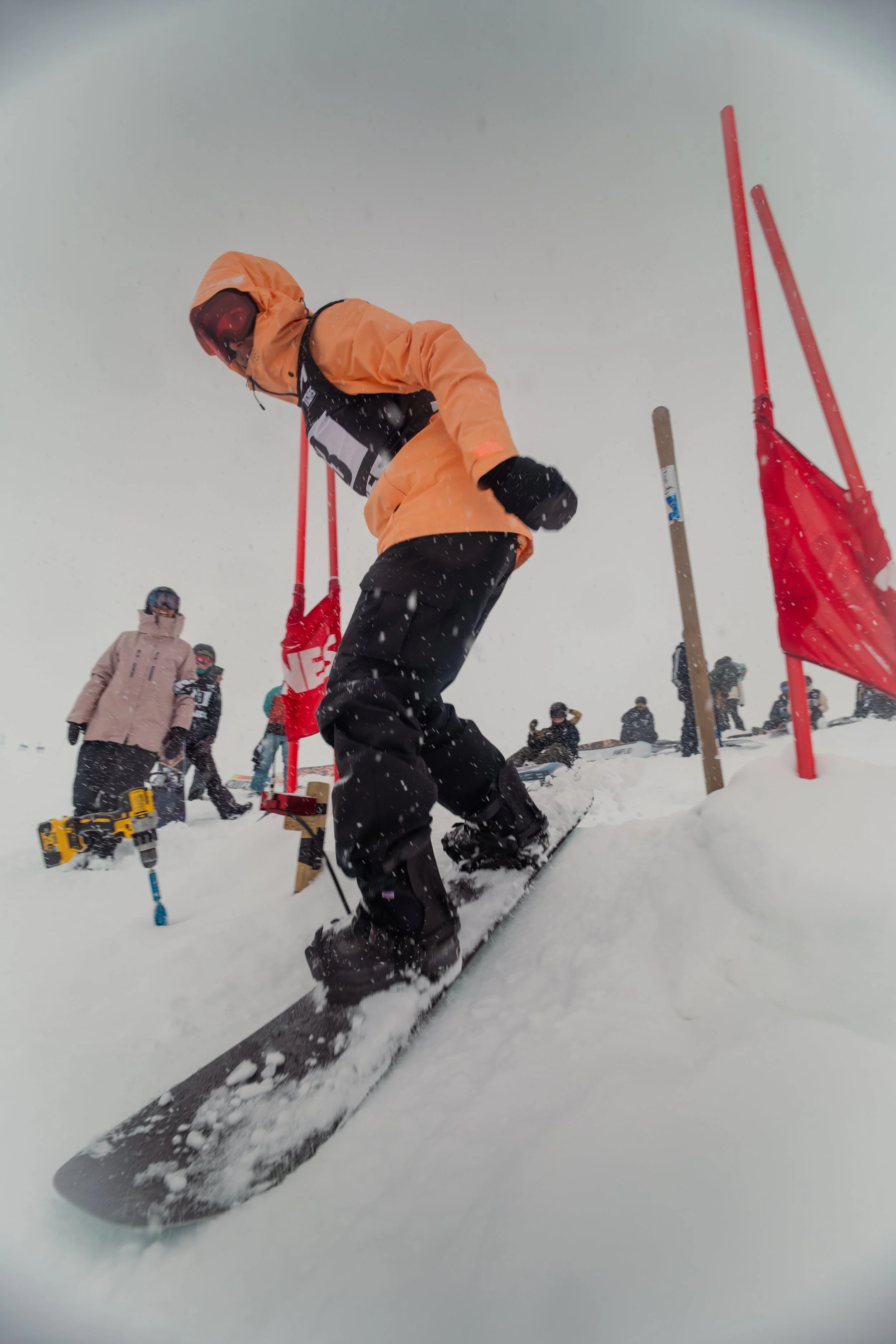 A snowboarder in an orange jacket and black pants riding down a snowy slope during a snowboarding event with other participants and officials in the background.
