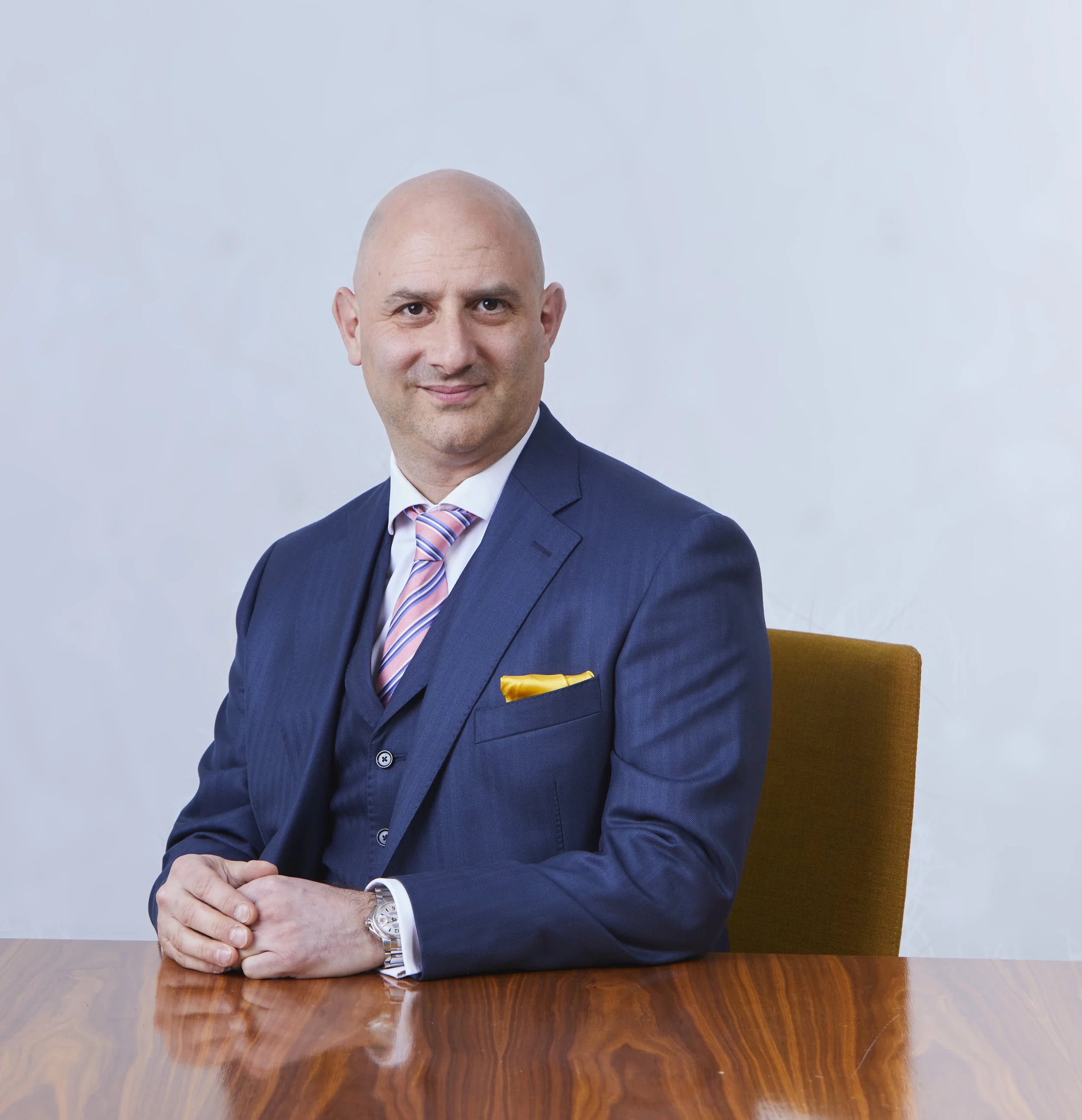 A bald man in a dark blue suit with a pink and blue striped tie and a yellow pocket square, sitting at a wooden table with a neutral background.