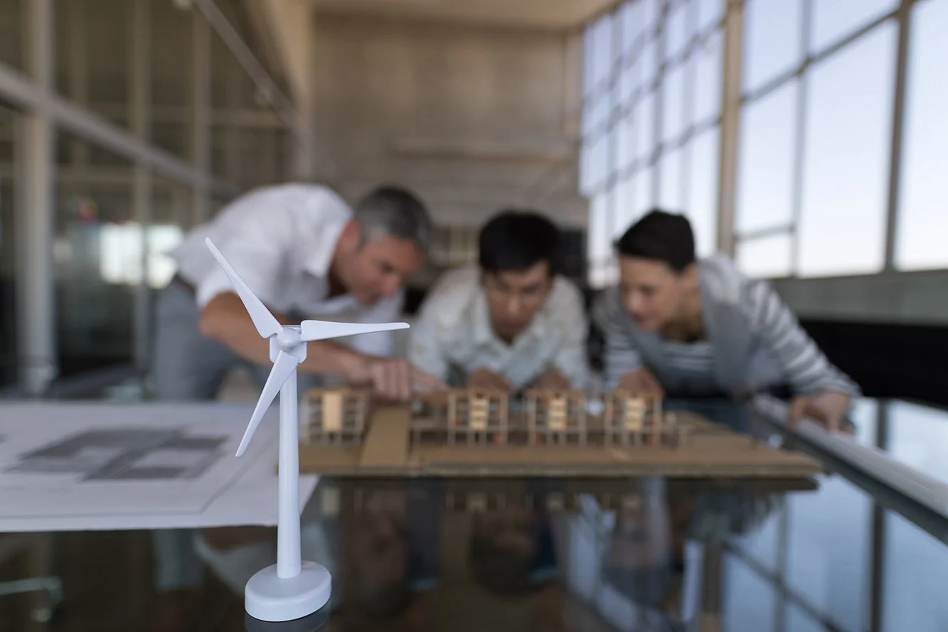 Small wind turbine model on table with three people examining architectural models in background.