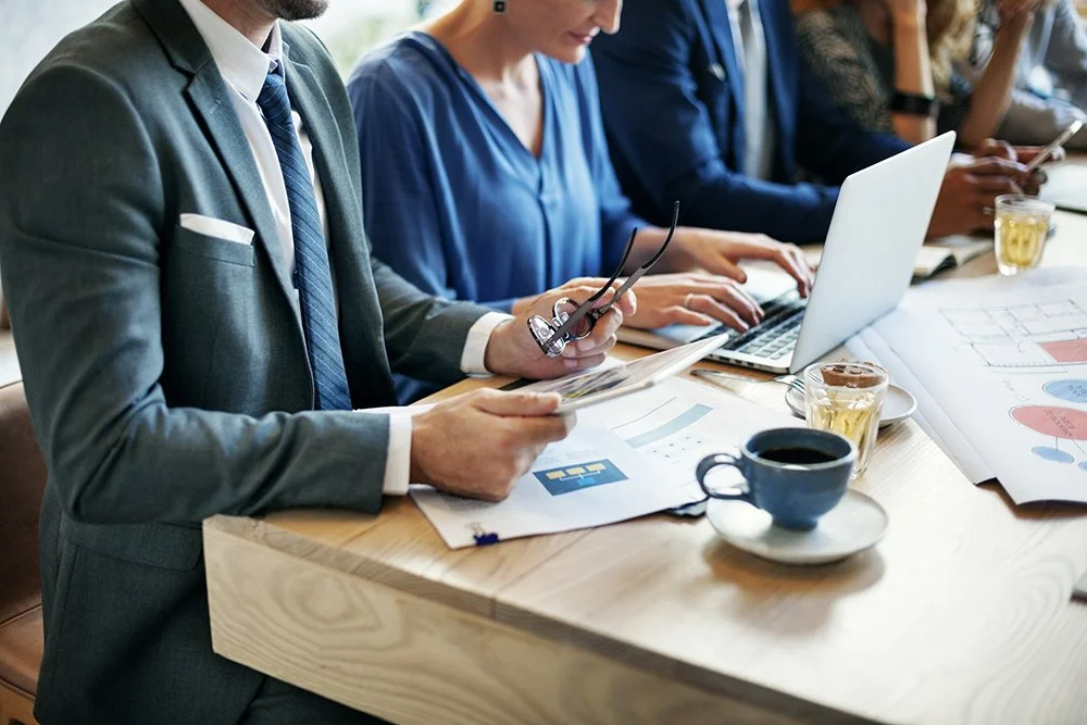 Business meeting with people working on laptops, reviewing documents, and engaging in discussion at a wooden table with coffee and drinks.