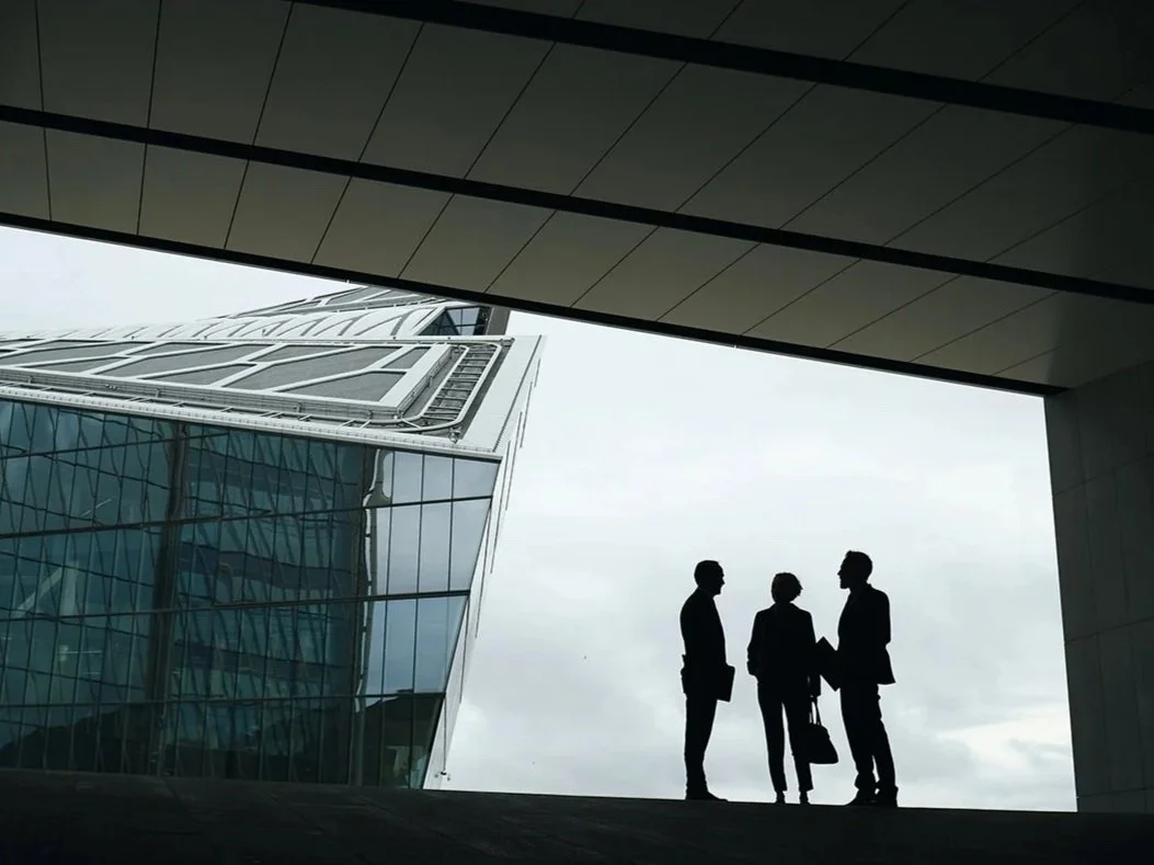 Silhouettes of four people standing and talking outside modern glass building under an overpass.