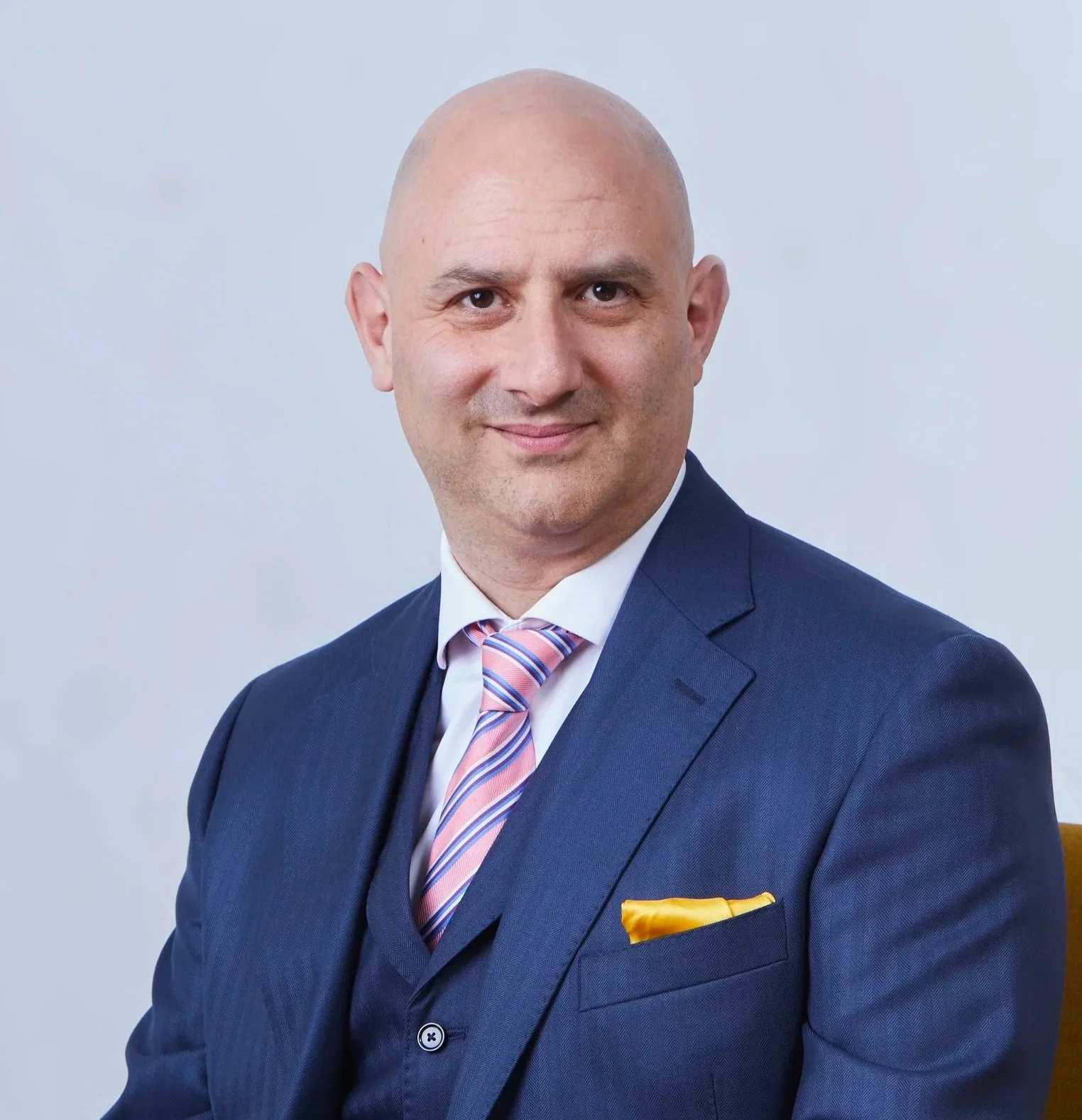 Taner Dedezade in a navy blue suit with a light pink striped tie sitting at a wooden table, smiling at the camera against a plain light gray background.