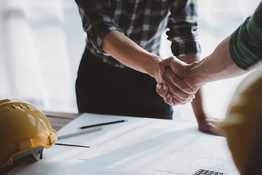 Two people shaking hands over a table with construction helmets and blueprints.