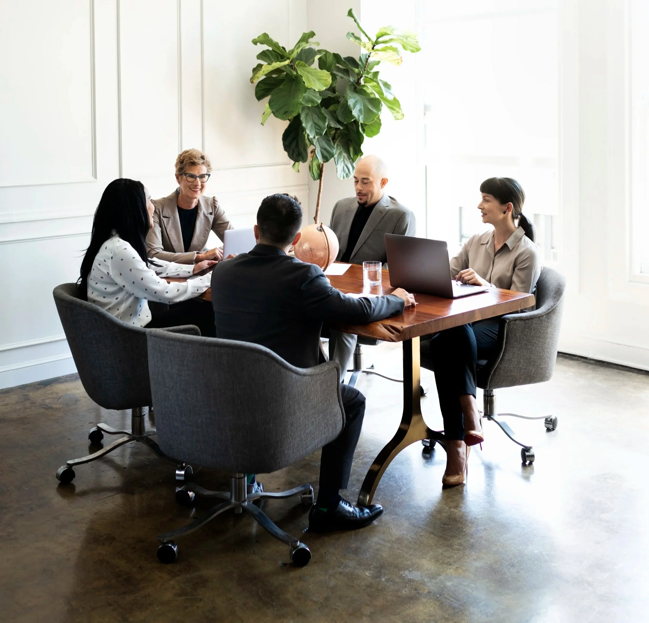 A diverse group of five business professionals in a meeting room, sitting around a wooden table with laptops, a globe, and glasses of water, engaged in discussion and smiling.