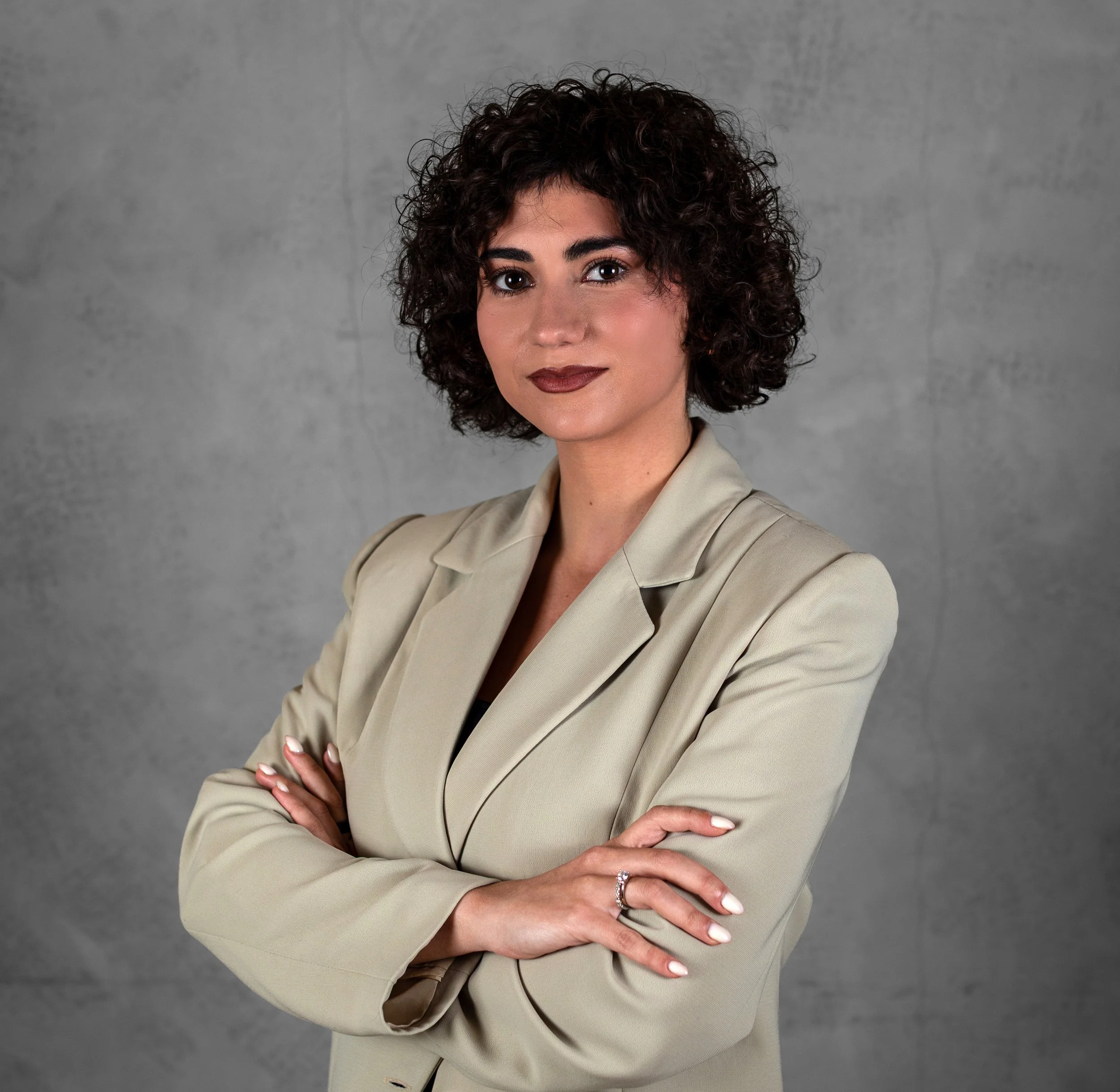 A woman with short curly dark hair, wearing a beige blazer, crosses her arms and faces the camera against a gray background.