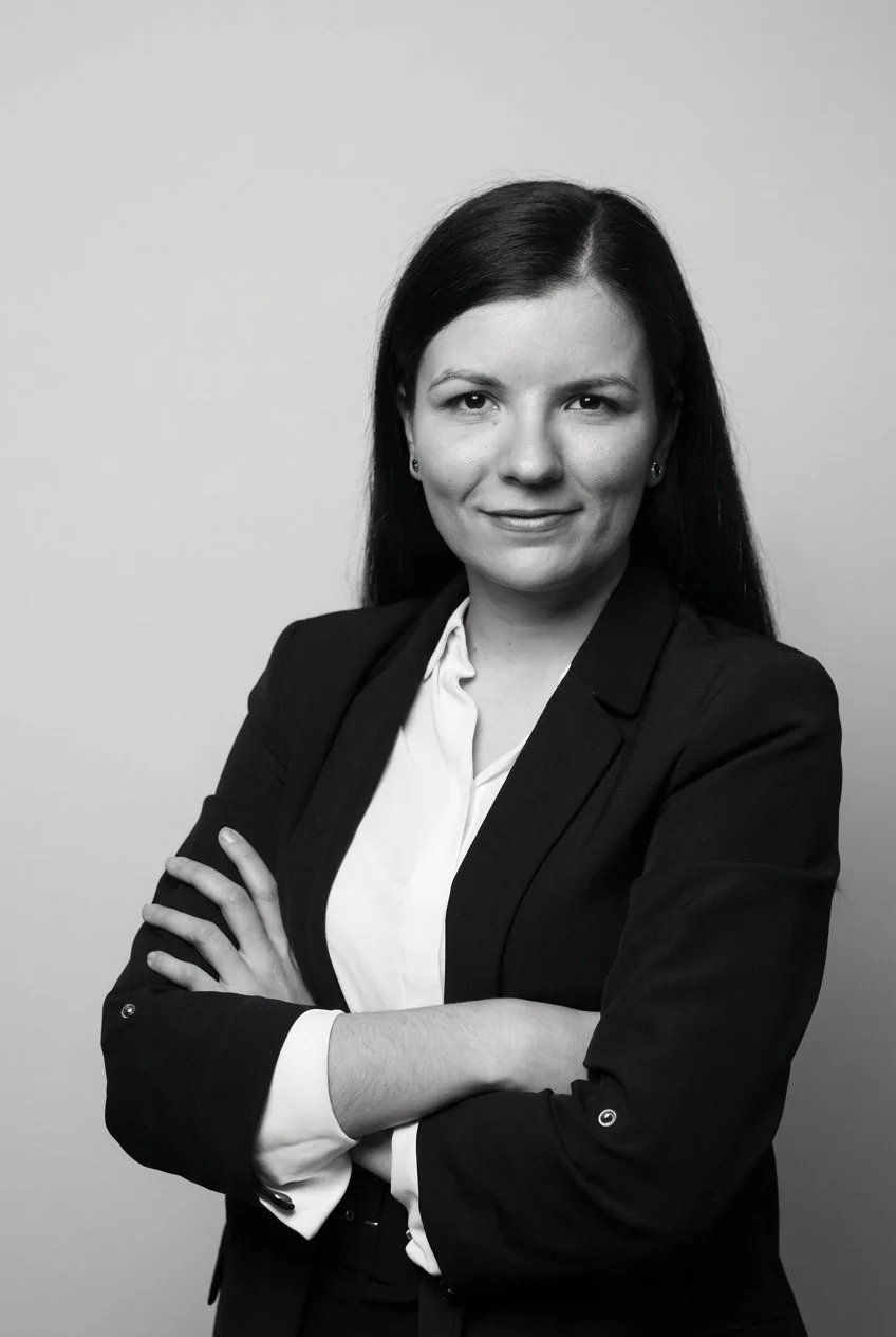 Black and white portrait of Zuzana Vysudilova​ in a business suit with arms crossed, smiling slightly, against a plain background.