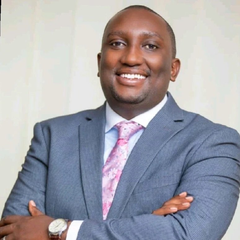 A smiling man in a gray suit with a pink and white patterned tie, arms crossed, standing against a plain background.