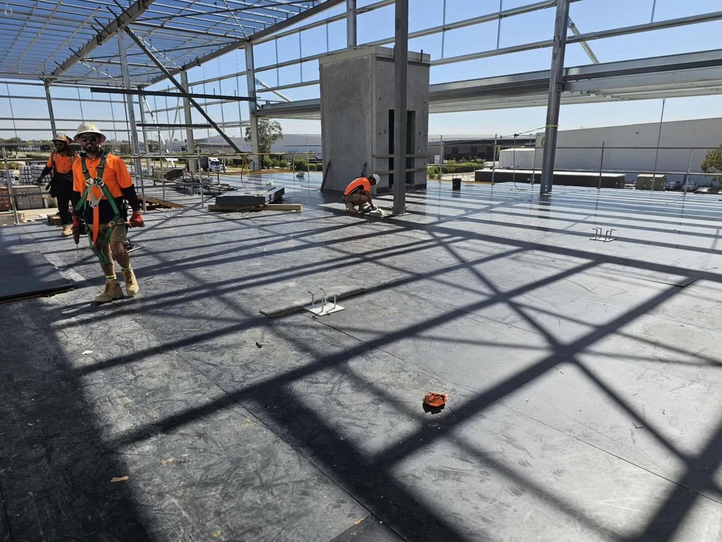 Construction workers wearing safety gear on a rooftop during daytime with sunlight and shadows of the roof structure on the floor.