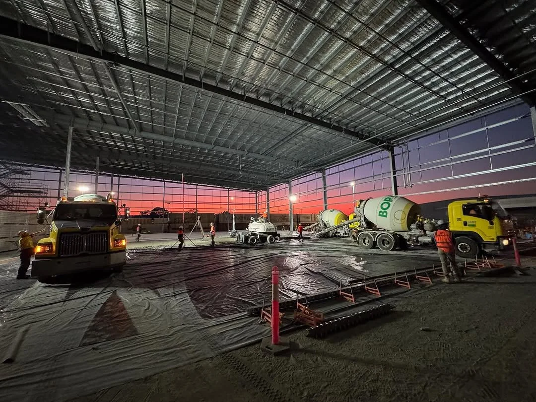 Construction workers pouring concrete inside a large building framework during sunset, with cement mixer trucks and construction equipment present.