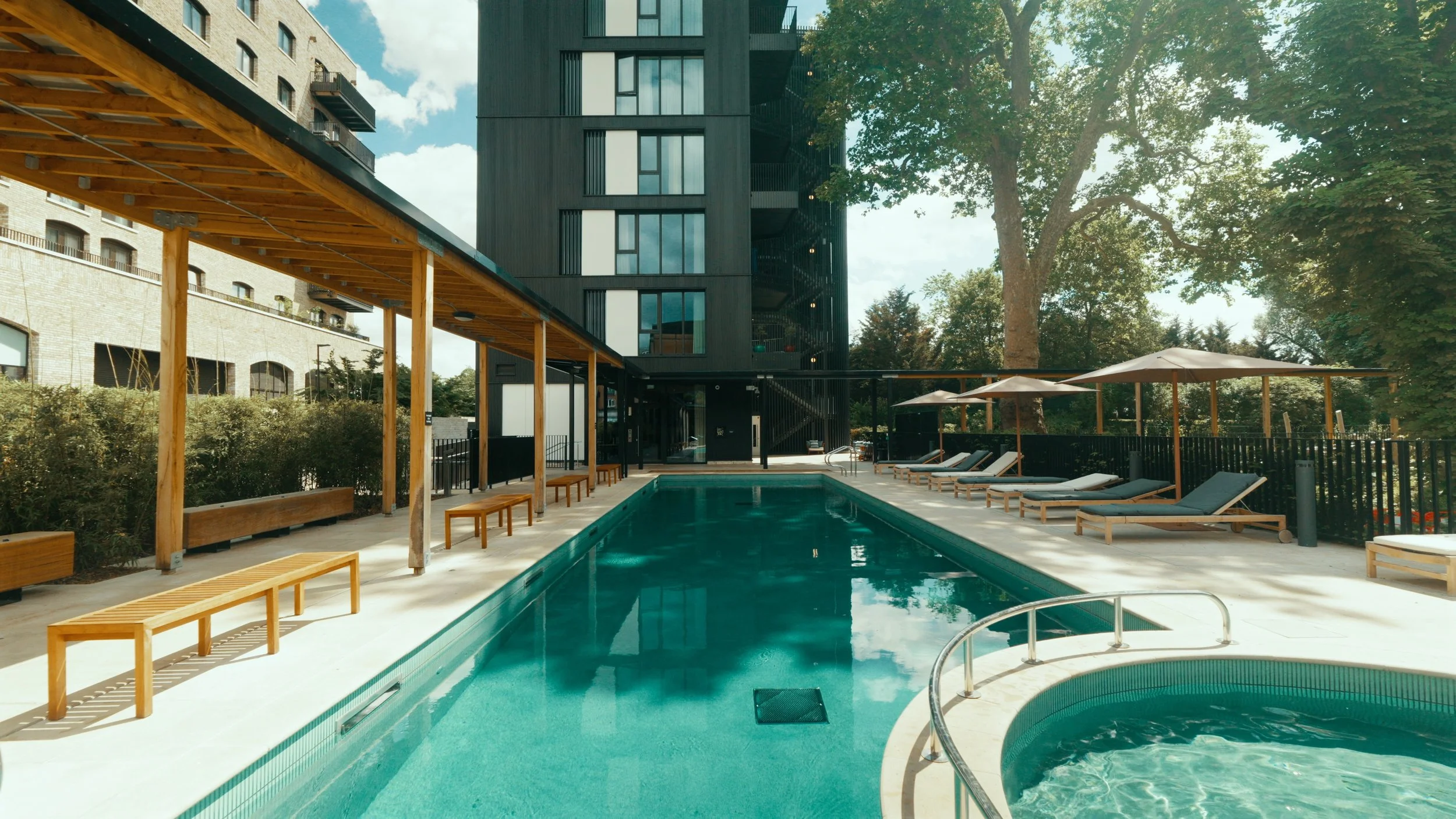 Outdoor swimming pool area at a modern apartment complex, with lounge chairs and shaded umbrellas, surrounded by trees and tall buildings.