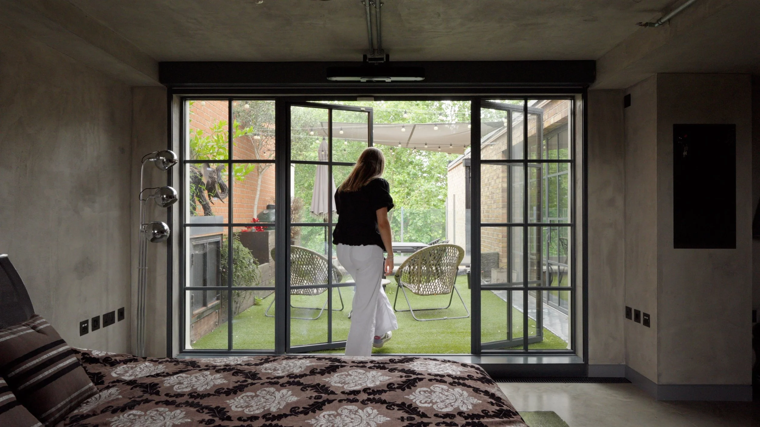 A woman in black top and white pants walking outside through glass doors to a patio with chairs and greenery