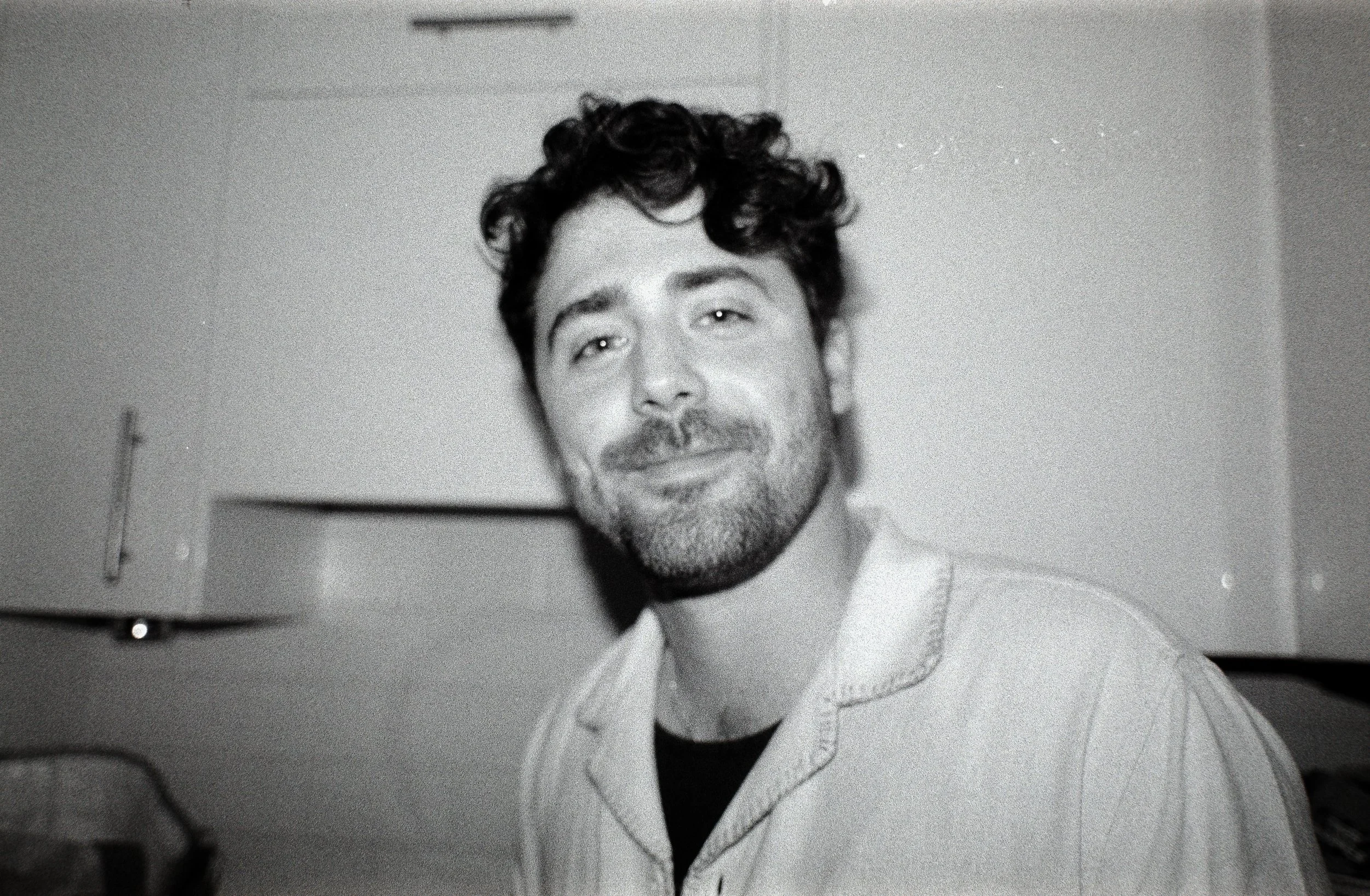Black and white photo of a young man with curly hair and a beard, smiling and looking at the camera, indoors.