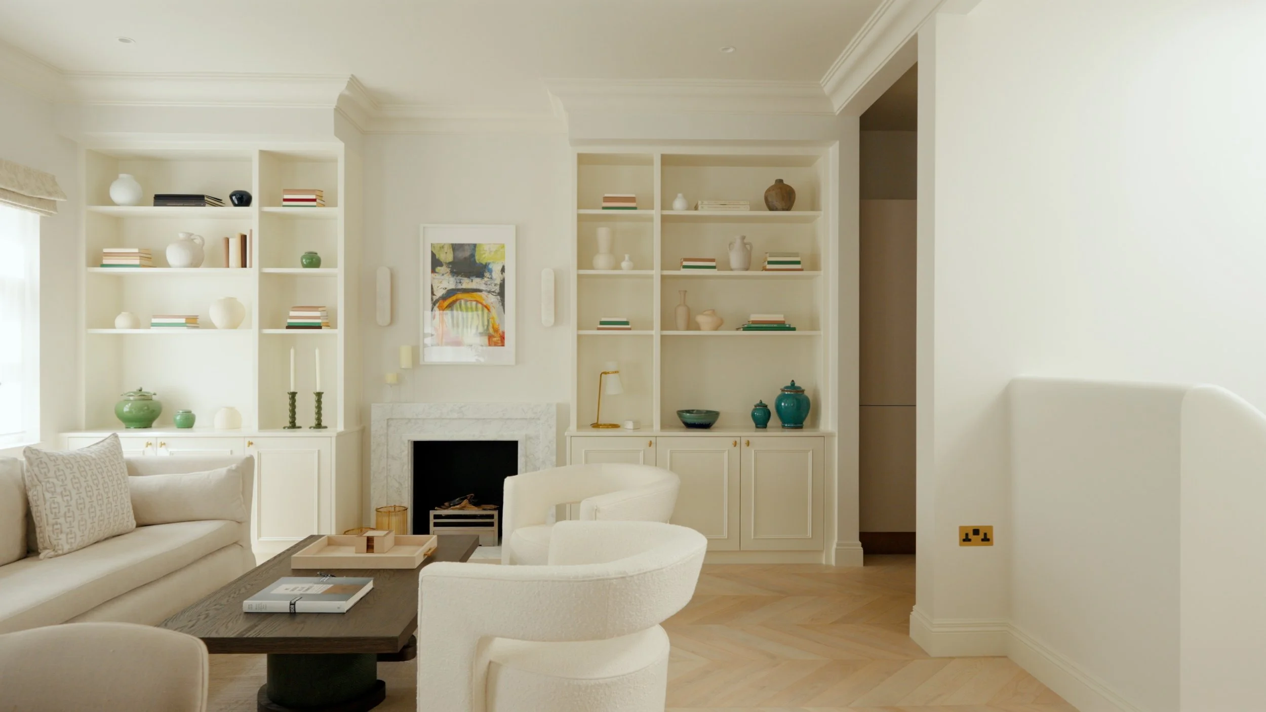 Living room with white walls, built-in shelves with decorative vases, books, and art, cream sofas, a dark wood coffee table, and a white marble fireplace.