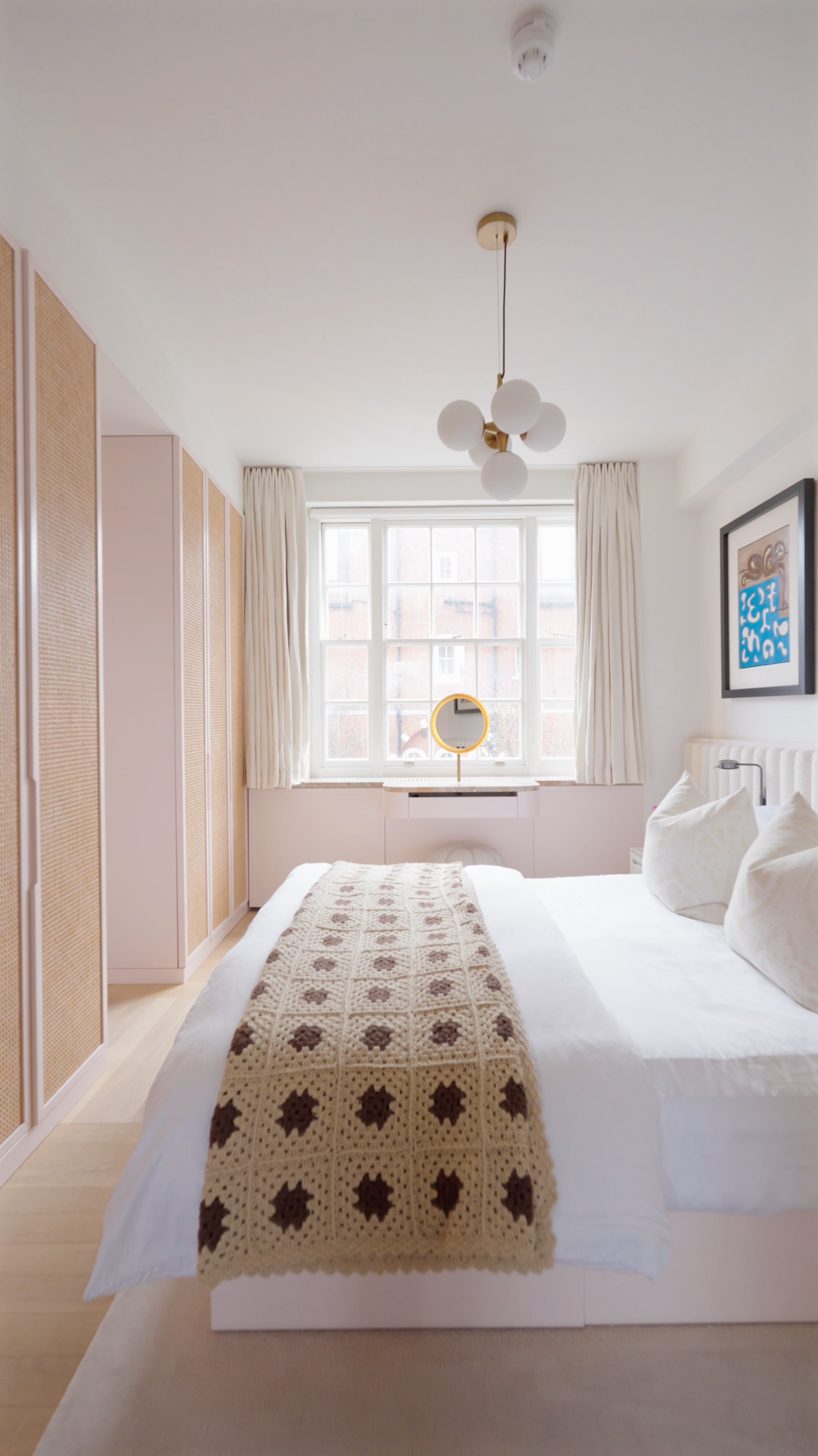 A neatly made bed with white bedding and pillows, a crocheted blanket with a brown floral pattern, positioned in a bright hotel room with a large window, white curtains, artwork on the walls, a modern ceiling light, and a circular mirror on the window ledge.