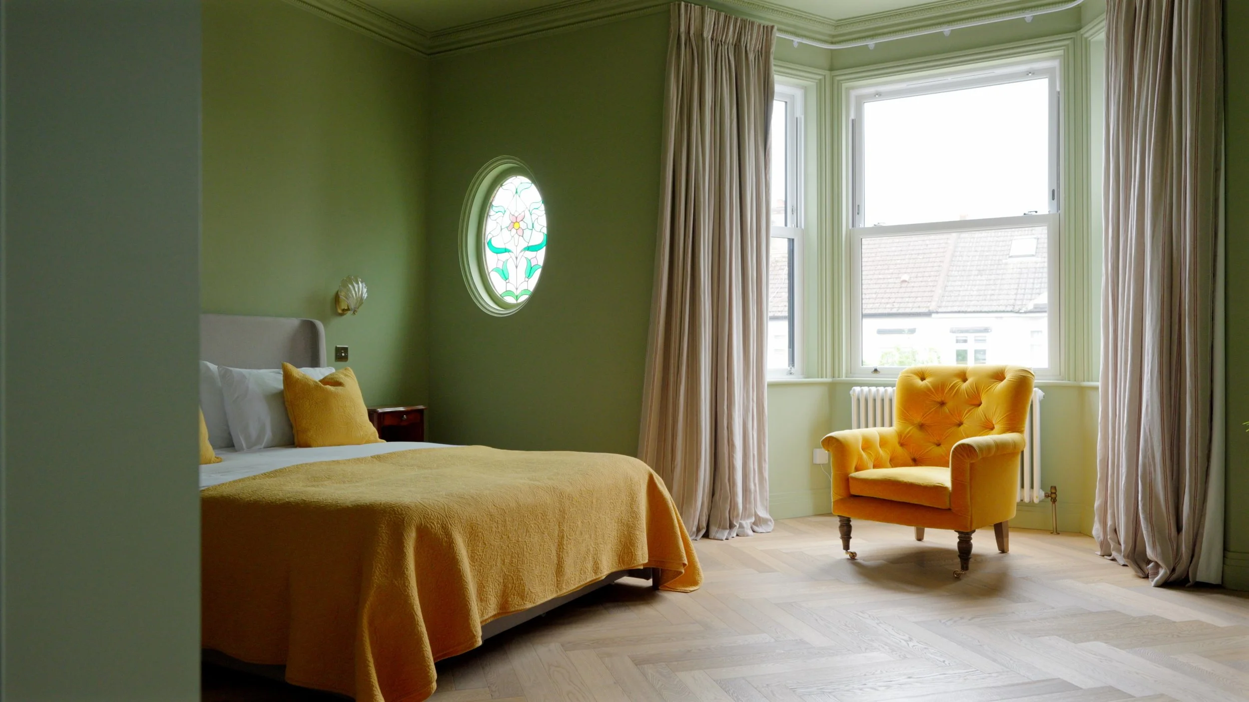 A cozy bedroom with green walls, a large bed with a yellow bedspread and pillows, a small side table, a yellow armchair, large windows with curtains, and a decorative stained glass window.