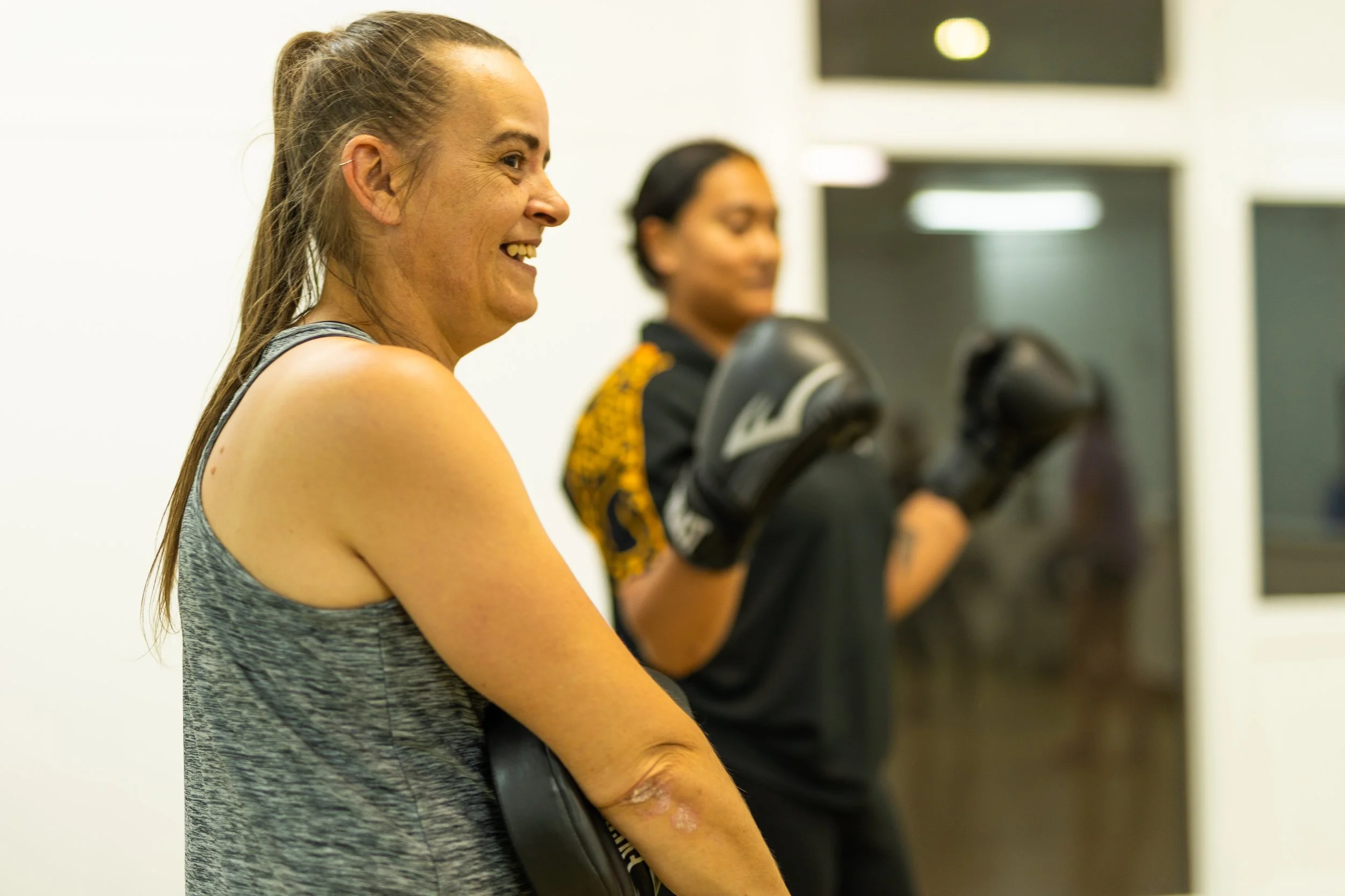 Two women in a gym, one in the foreground with a smile, wearing a gray tank top, and the other in the background with boxing gloves, wearing a black shirt with yellow accents.