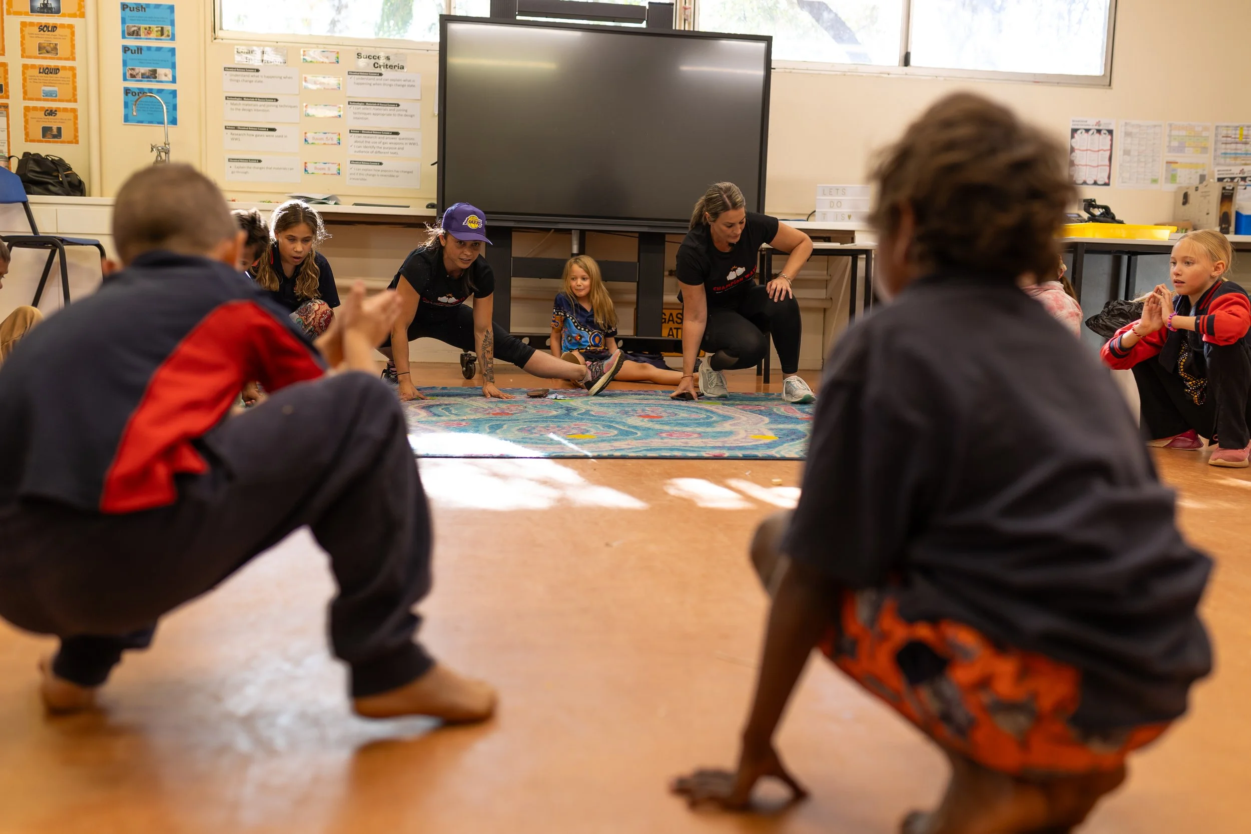 Children and teachers on the floor in a classroom, engaged in a stretching or movement activity, with a large screen and educational posters on the wall.