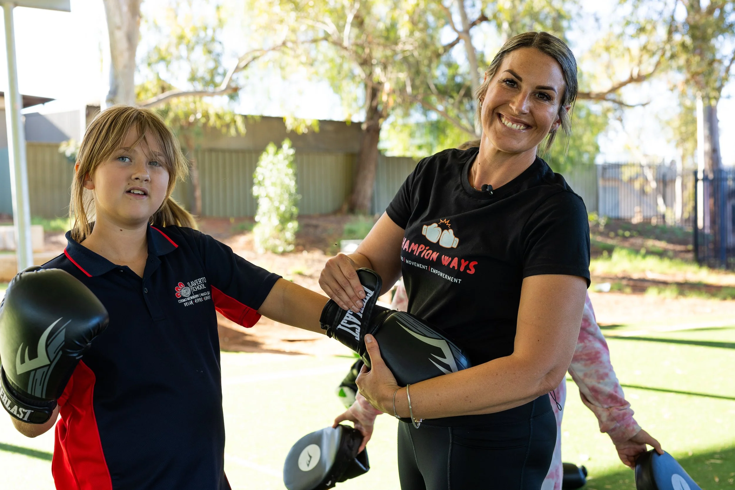 A woman and a young girl are outdoors, with the girl wearing boxing gloves and the woman smiling while holding boxing pads. The woman is wearing a black T-shirt with the words 'Champion Ways' printed on it, and the girl is wearing a dark polo shirt with a logo. They are surrounded by trees and a fence in the background.