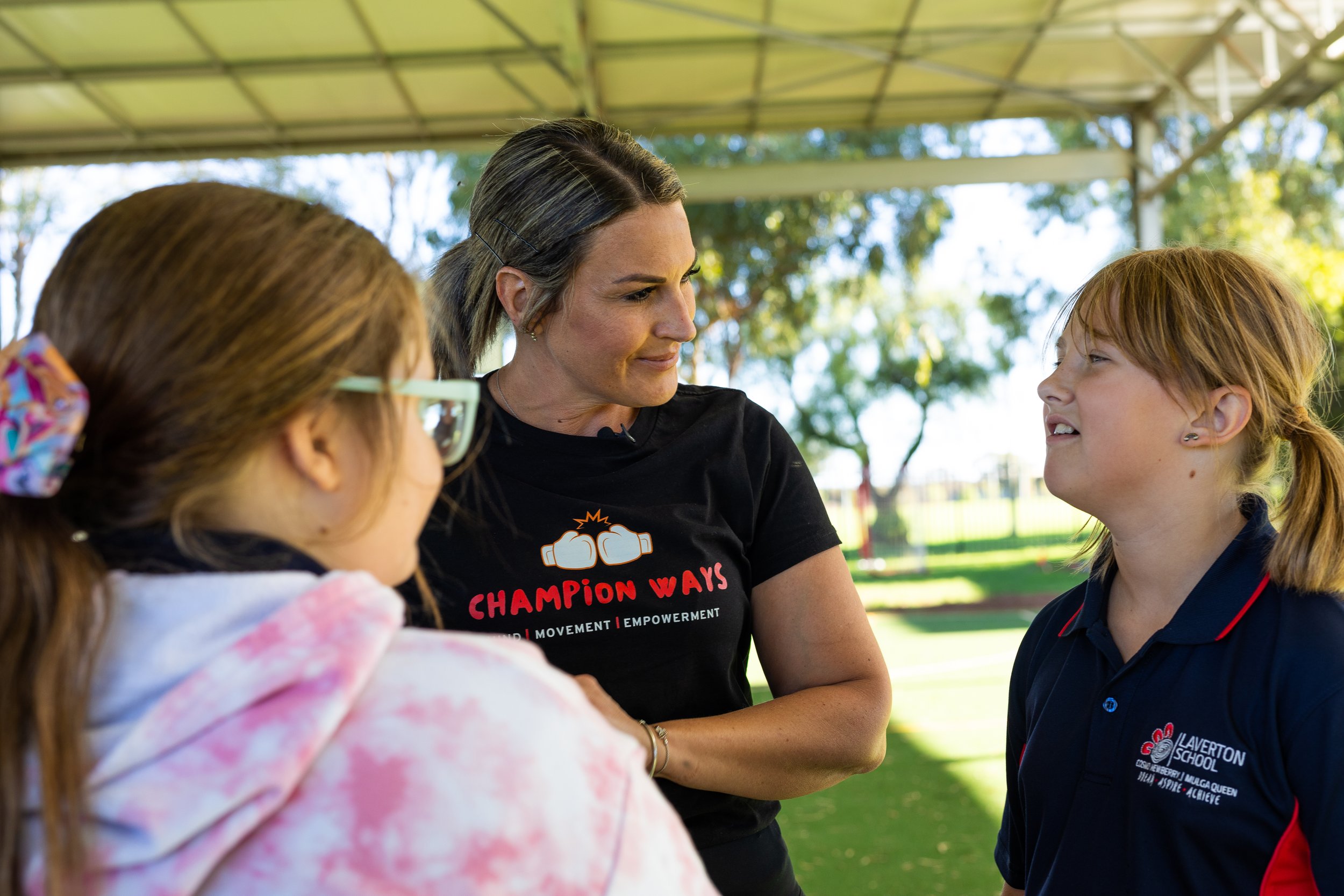 A woman and two girls standing outdoors under a covered structure, engaging in a conversation. The woman is wearing a black T-shirt with the words 'Champion Ways' and an illustration of two boxing gloves. One girl with glasses and a pink hoodie is facing the woman, while another girl in a navy blue polo shirt with a logo is smiling at the woman. There are trees and a grassy field in the background.
