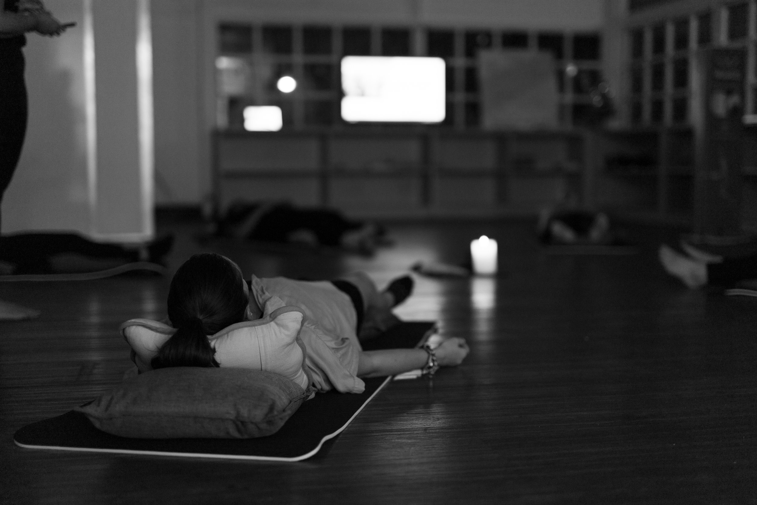 A woman lying on her side on a yoga mat in a dimly lit room, with her head resting on a pillow, participating in a meditation or relaxation session, with several others lying on mats in the background and a candle and TV screen in the room.