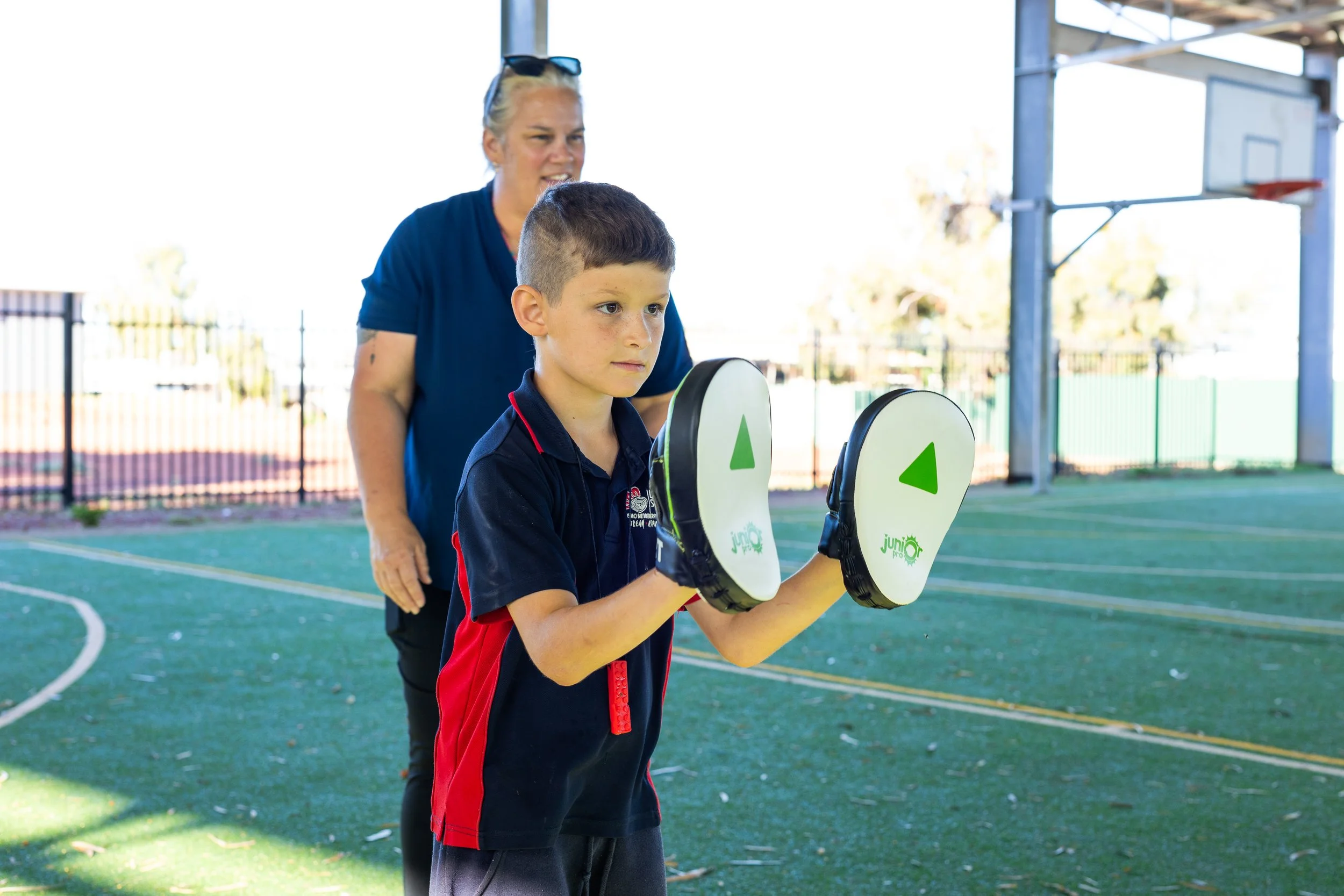 A young boy practicing boxing on an outdoor court, supervised by a woman standing behind him.