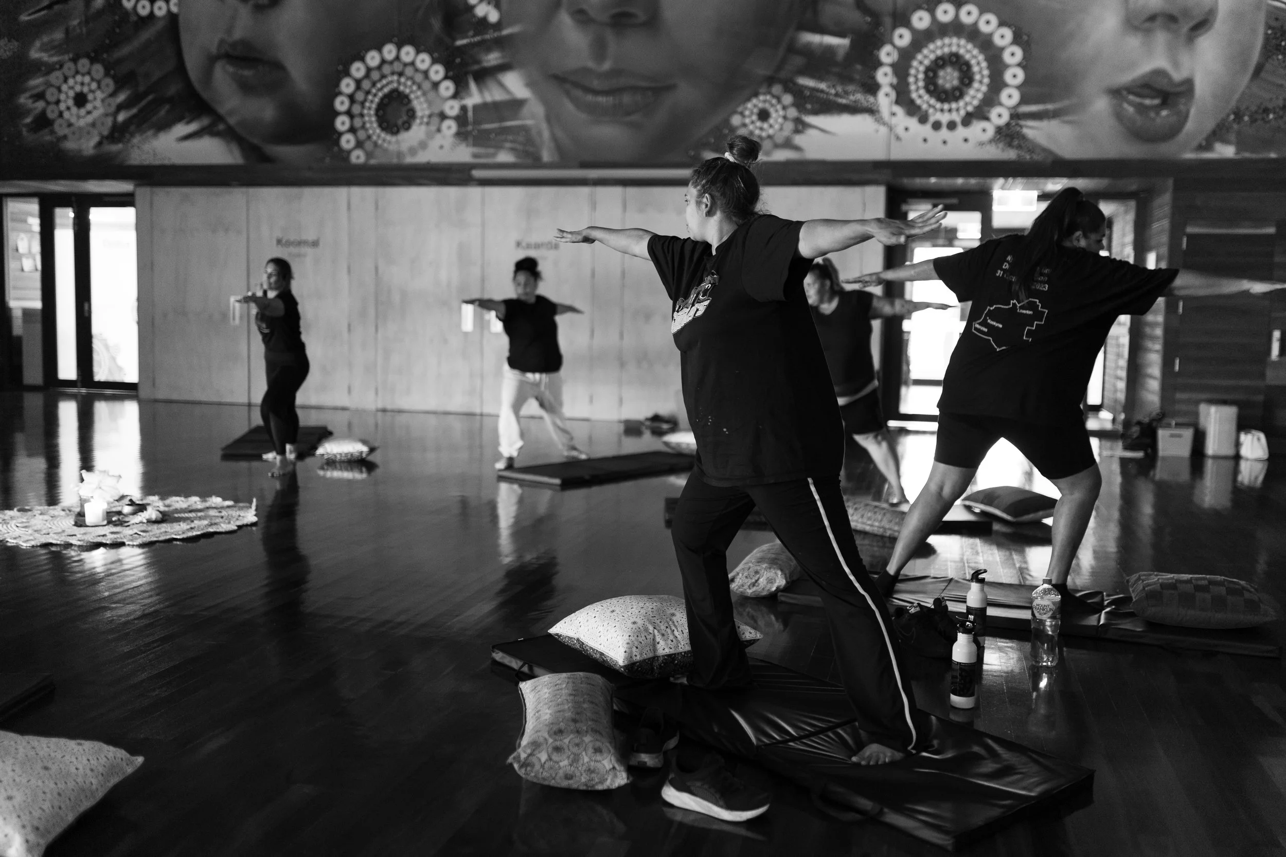 People participating in a yoga class practicing poses on mats in a studio with wooden walls and colorful artwork on the wall.