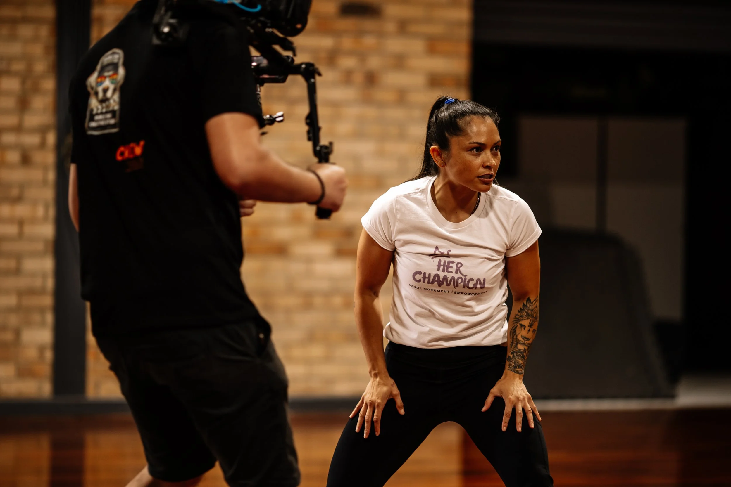 A woman in a white T-shirt and black pants is in a fitness class, standing with hands on her thighs, talking to a man in black clothes and holding a camera.