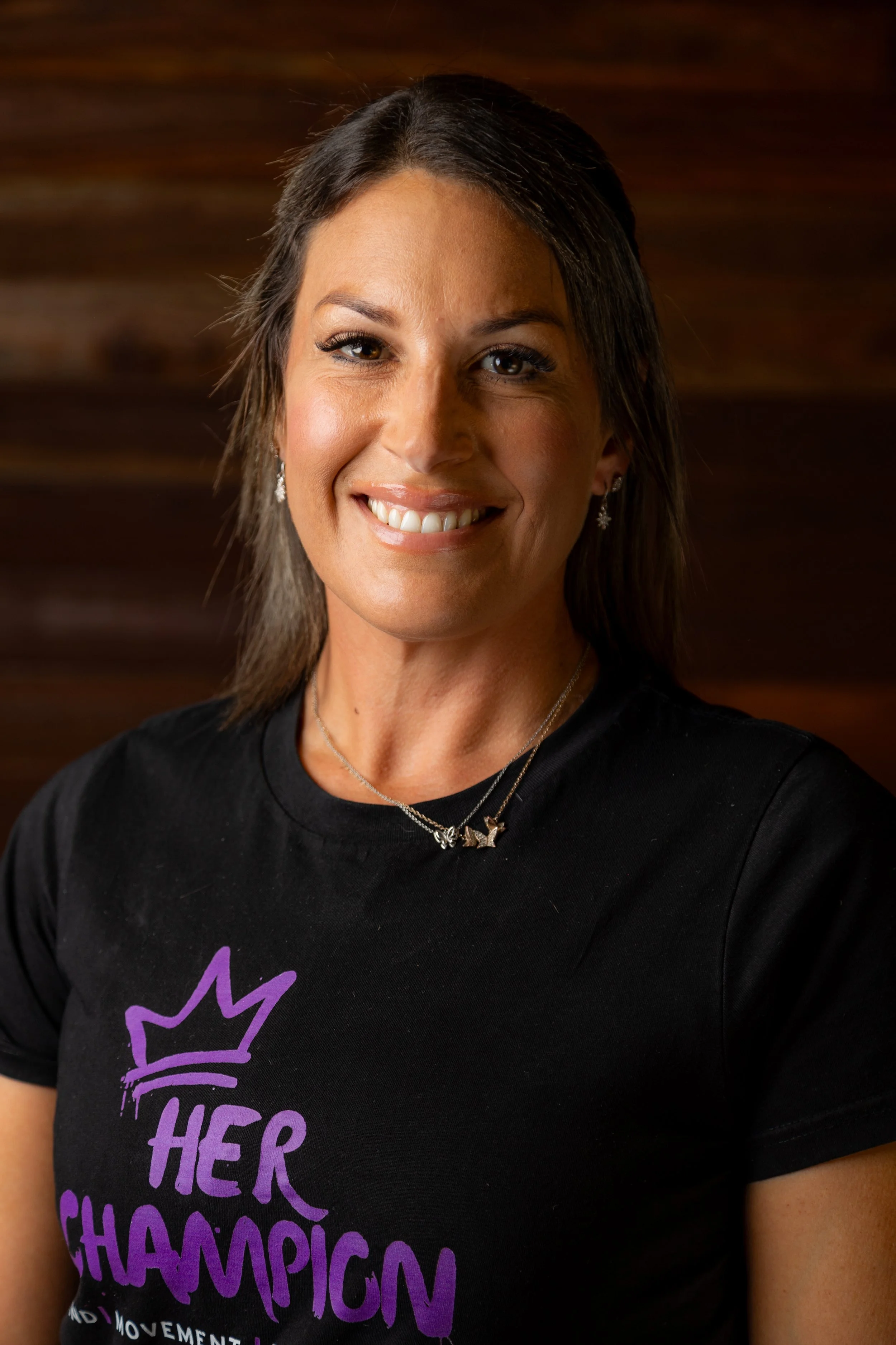 Close-up of a woman smiling, wearing a black T-shirt with purple text that reads 'Her Champion' and a crown logo, with a wooden background.