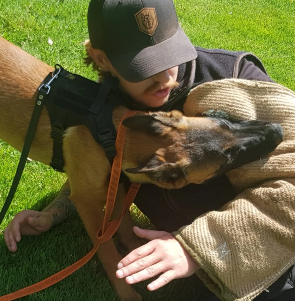 A man wearing a black and gray hat and a black jacket is lying on the grass, hugging a brown dog. The dog, which appears to be a Belgian Malinois, has a black face and ears, and is lying comfortably with its head resting on the man's chest. The man gently holds the dog's body, and they seem to be sharing an affectionate moment outdoors on a sunny day.