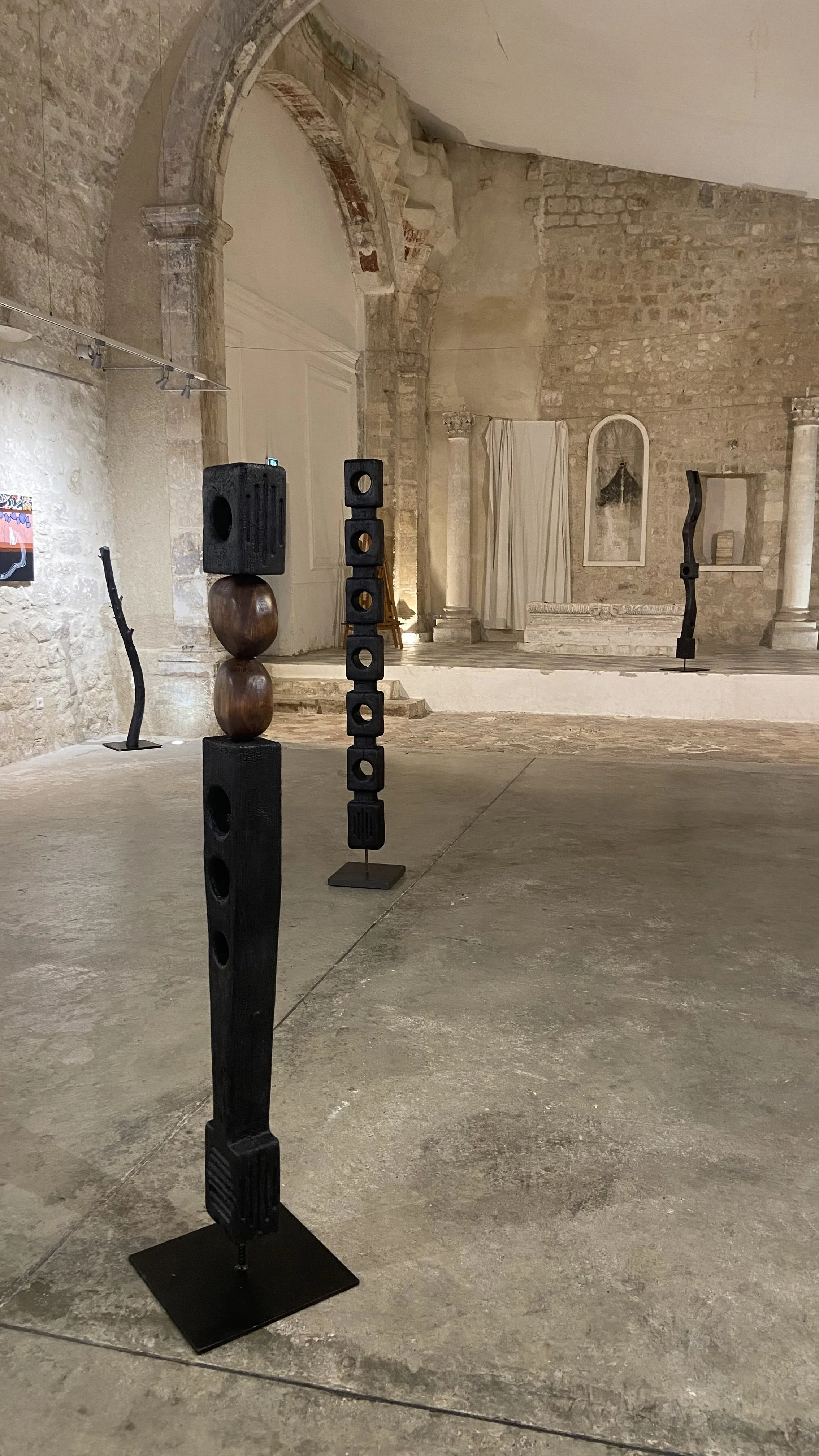 Une exposition d'œuvres d'art ou de sculptures dans une salle aux murs en pierre, avec des sculptures en bois et en métal disposées sur le sol.