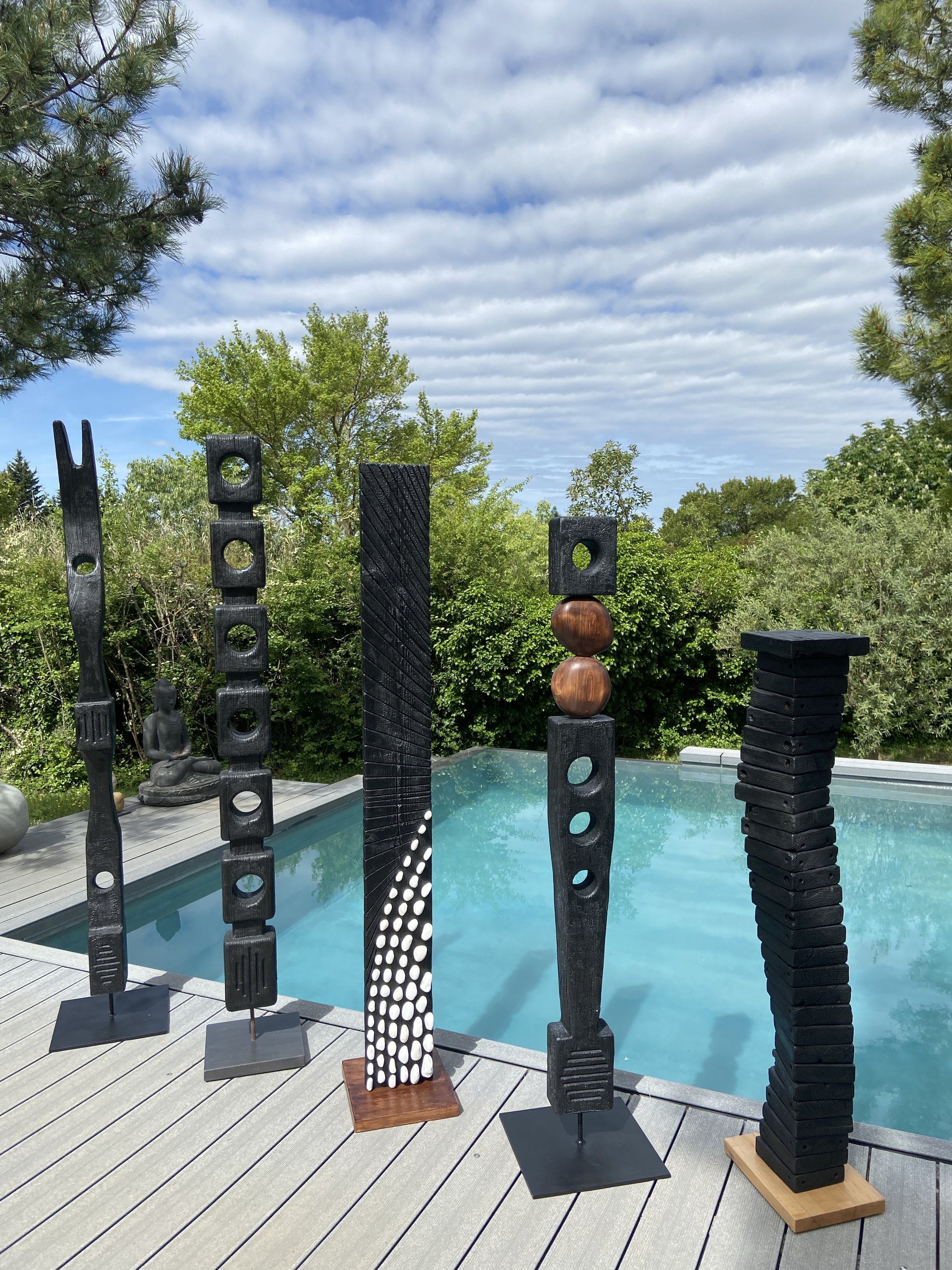 Sculptures en bois modernes de différentes formes et tailles, disposées sur une terrasse en bois près d'une piscine, avec des arbres et un ciel nuageux en arrière-plan.