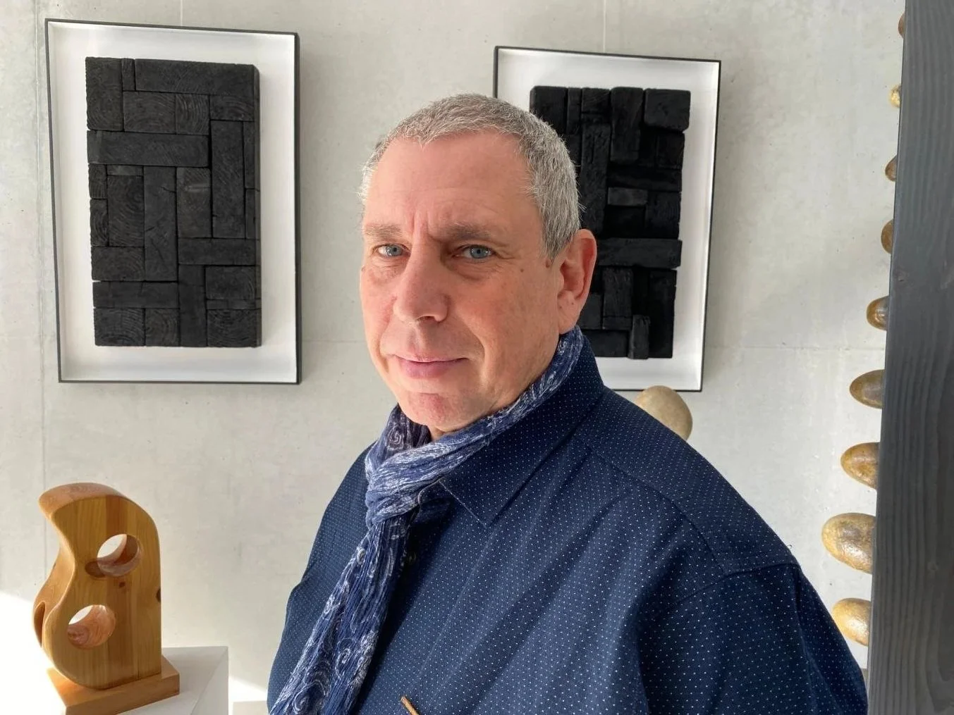 Un homme avec des cheveux gris, portant une veste bleue et une écharpe bleue, se tenant devant un mur décoratif avec deux tableaux en relief noirs et quelques sculptures en forme de pierres.