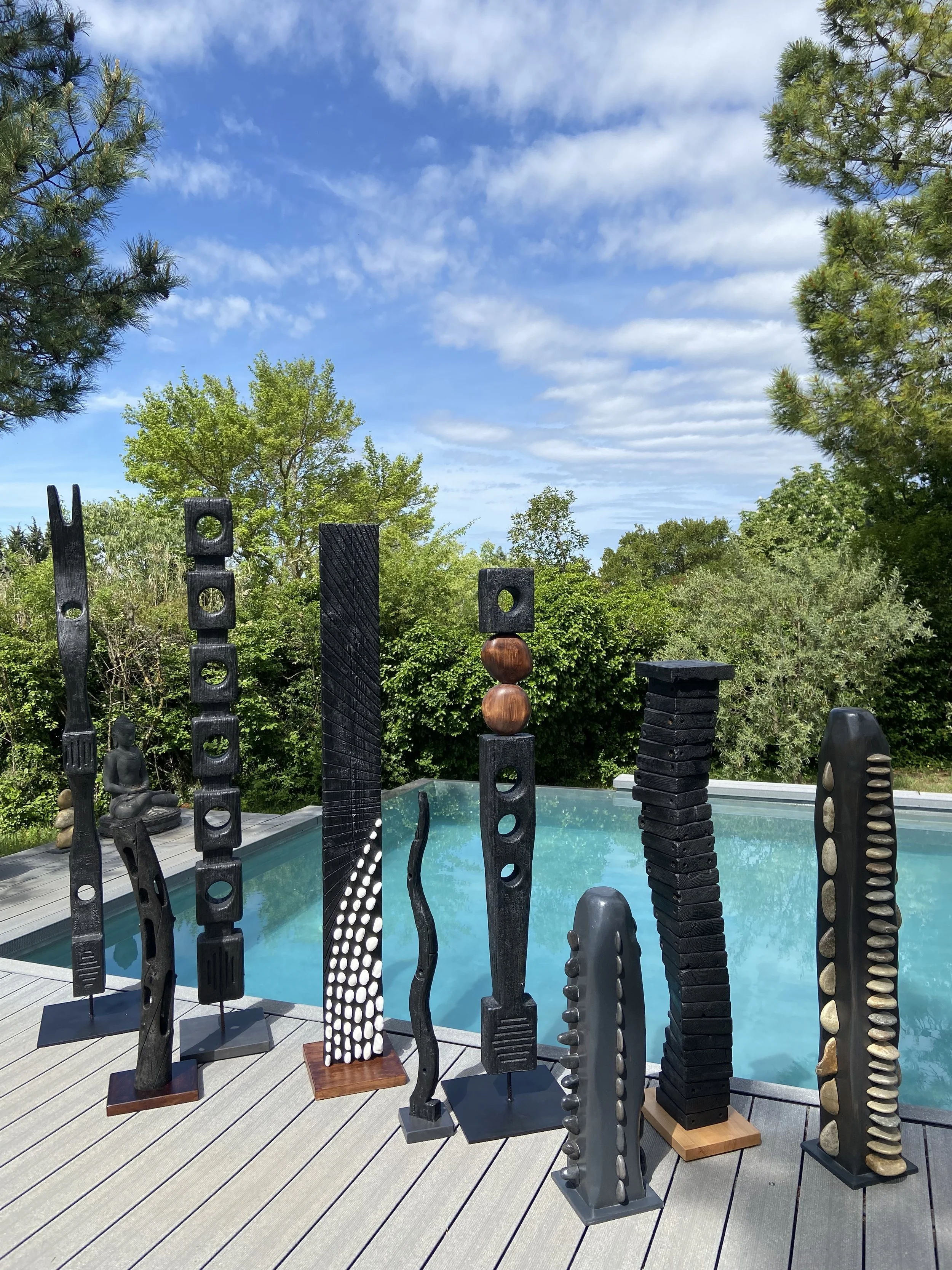 Sculptures modernes en bois noir et pierre, disposés sur une terrasse près d'une piscine, avec des arbres et un ciel nuageux en arrière-plan.