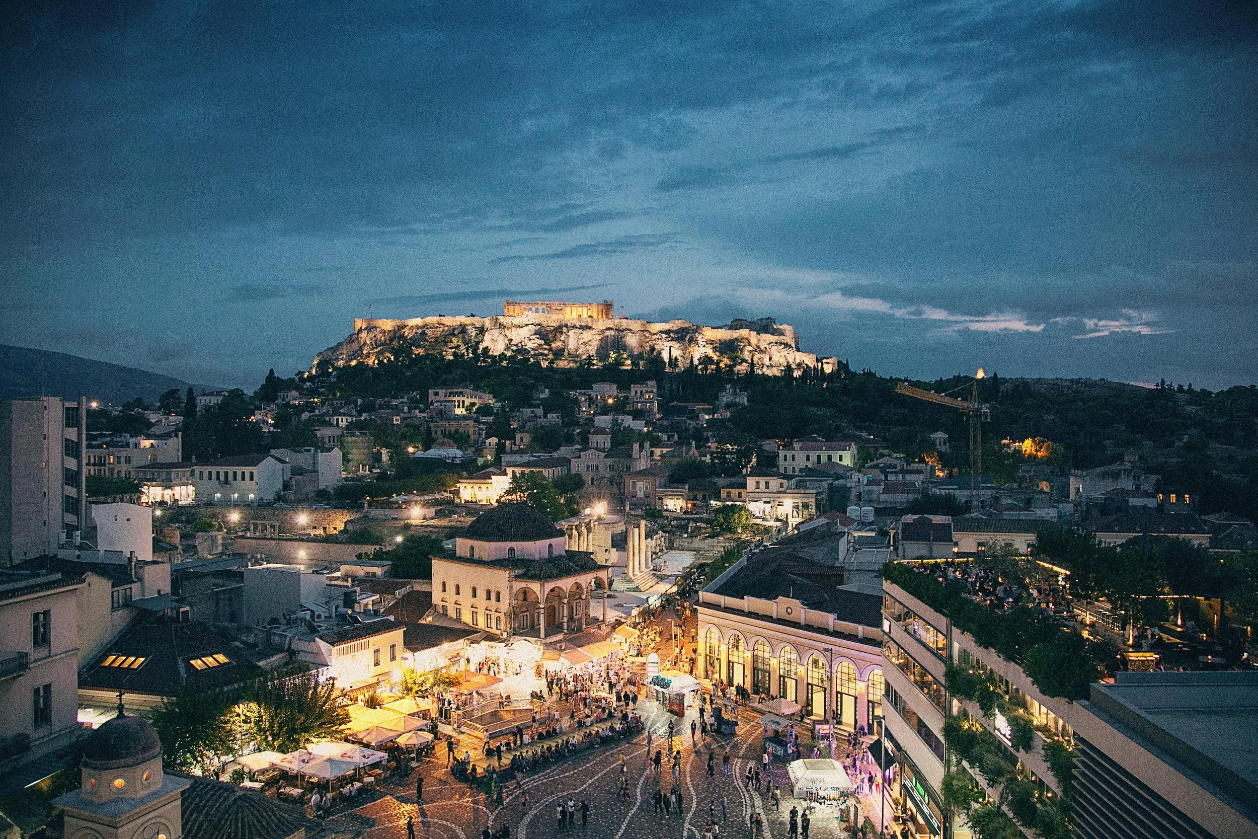 Abendansicht der Akropolis auf einem Hügel in Athen, geschmückt mit Beleuchtung, mit Stadt und Menschenmenge im Vordergrund.