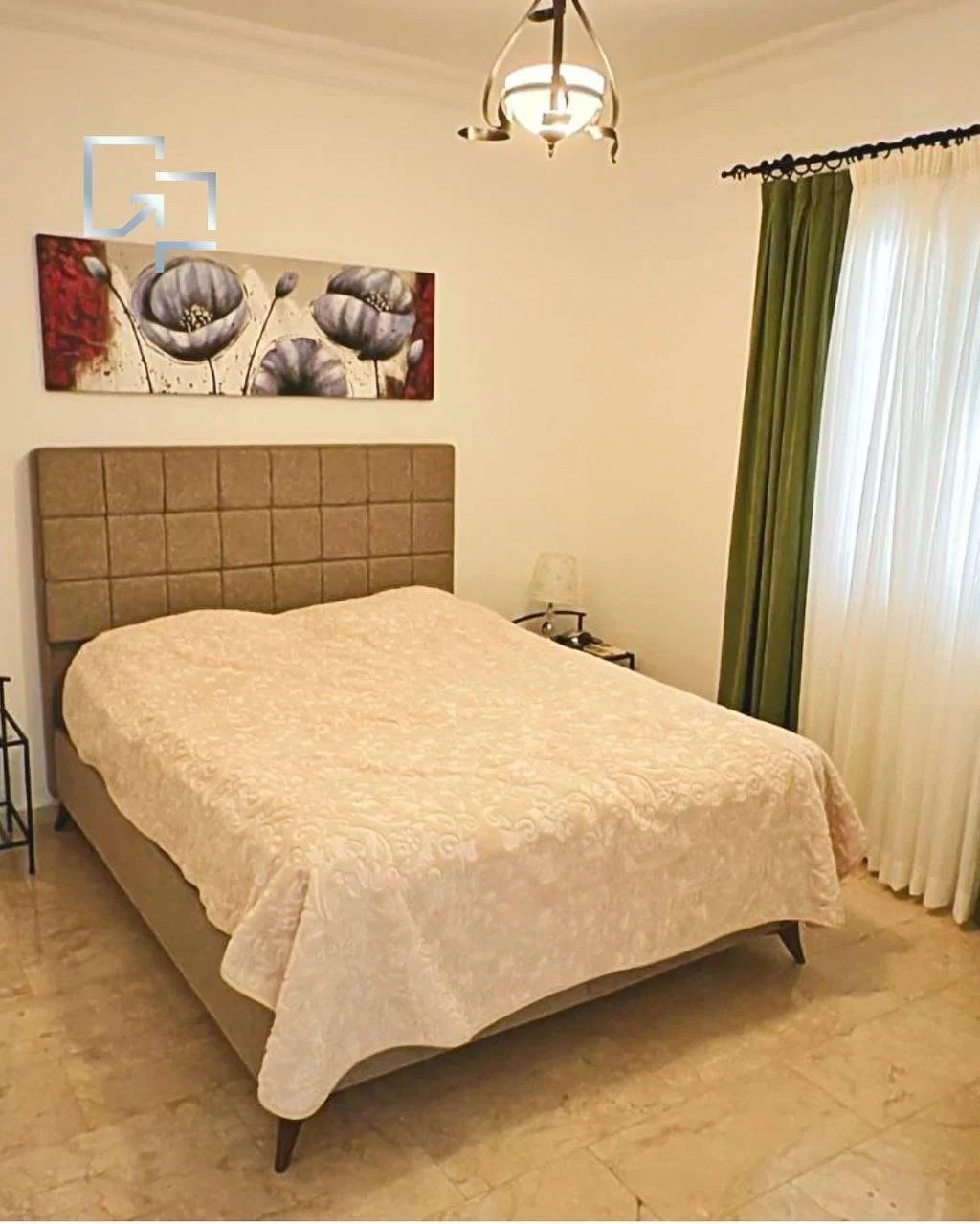 Ein Schlafzimmer mit einem großen Bett, einem beige Kopfteil, einer bedruckten Bettdecke, einem Nachttisch mit Lampe, einer Wanddekoration mit schwarzen Blumen, einem Fenster mit weißen Vorhängen und grünen Gardinen, sowie einem Deckenleuchter.