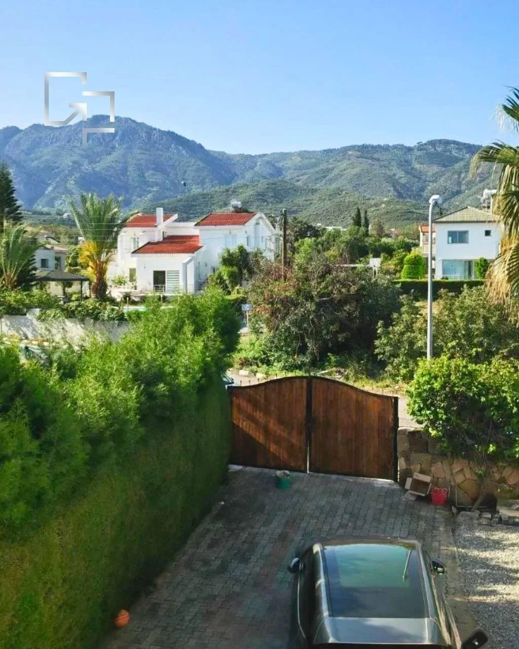 Ein Blick auf eine Hausterrasse mit einer großen Einfahrt, einem schwarzen Auto, einer hohen Hecke, einem Holztor, und im Hintergrund grüne Hügel unter einem blauen Himmel.