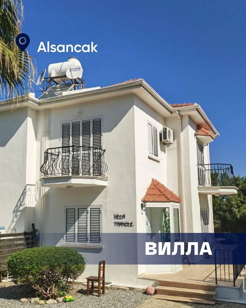 Mehrstöckige Villa mit weißen Wänden, kleinen Balkonen mit schwarzen Geländern, einem Dach mit roten Ziegeln, und einem Satelliten auf dem Dach, unter einem klaren blauen Himmel.