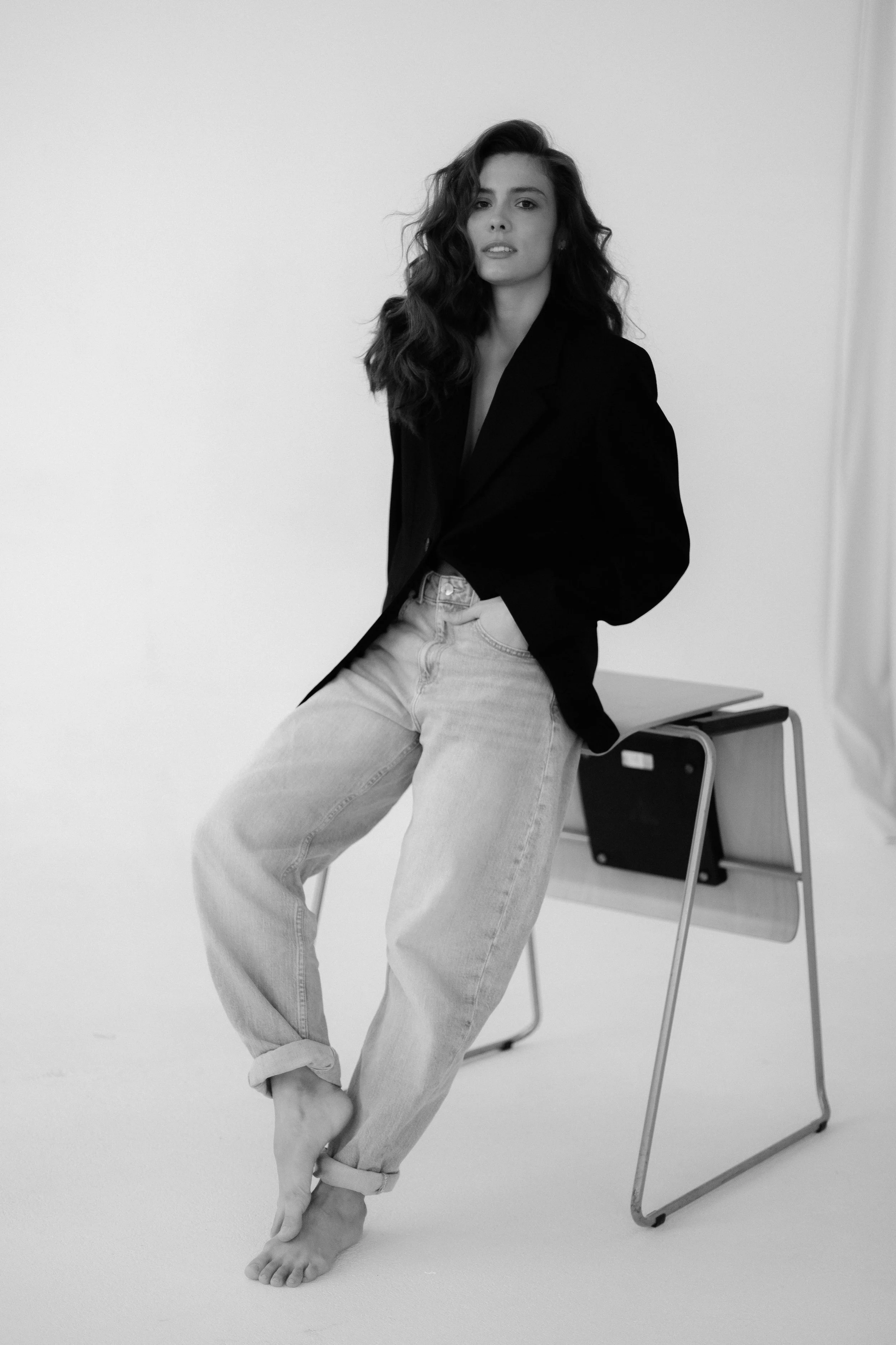 A woman with long, wavy hair wearing a dark blazer and light jeans, standing barefoot next to a desk in a minimalistic room.