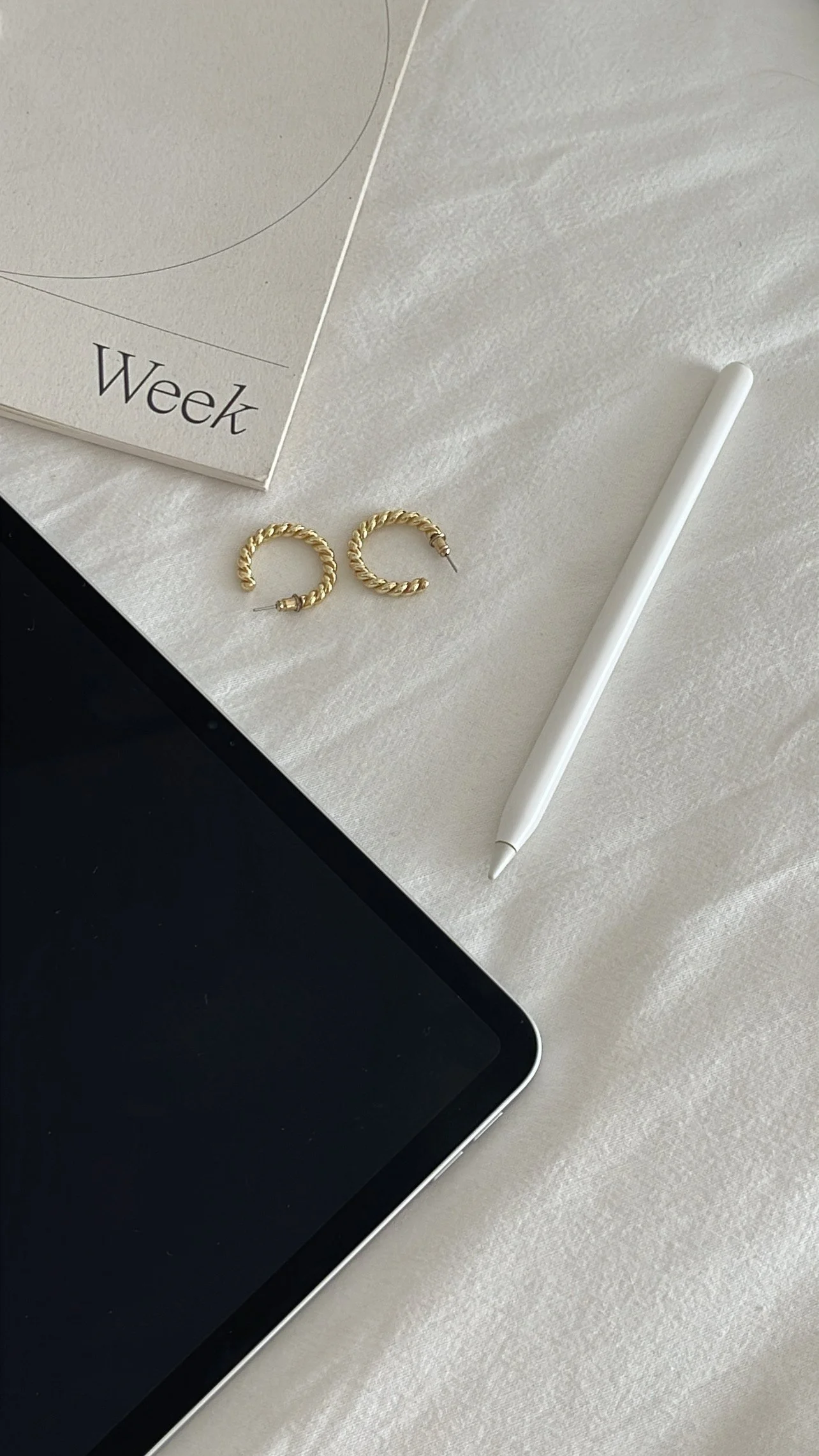 Flat lay of a workspace with a digital tablet, white stylus, gold hoop earrings, and a weekly planner on a textured white surface.