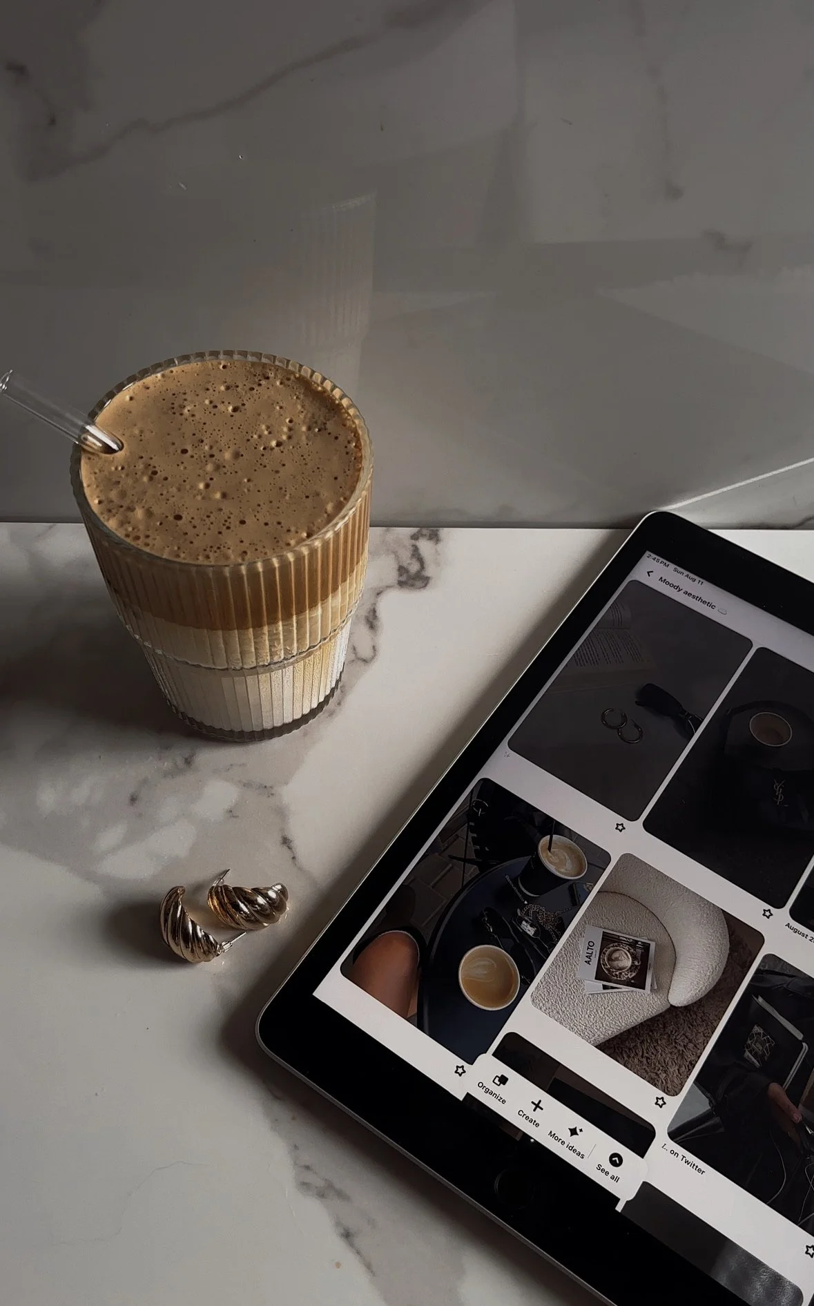 A tall glass of iced coffee with a straw on a white marble table next to a tablet displaying a photo gallery and a pair of silver hoop earrings.