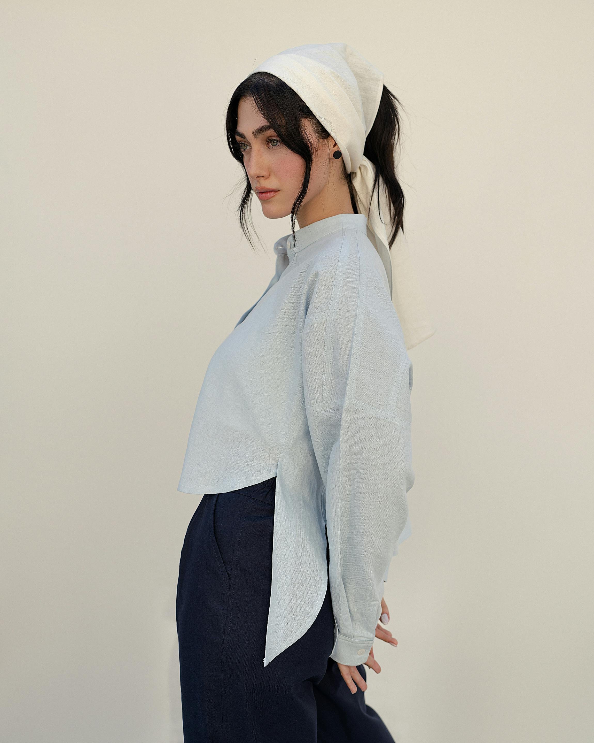 A woman with black hair and a white headscarf, wearing a light blue button-up shirt and dark pants, standing against a plain light-colored background.