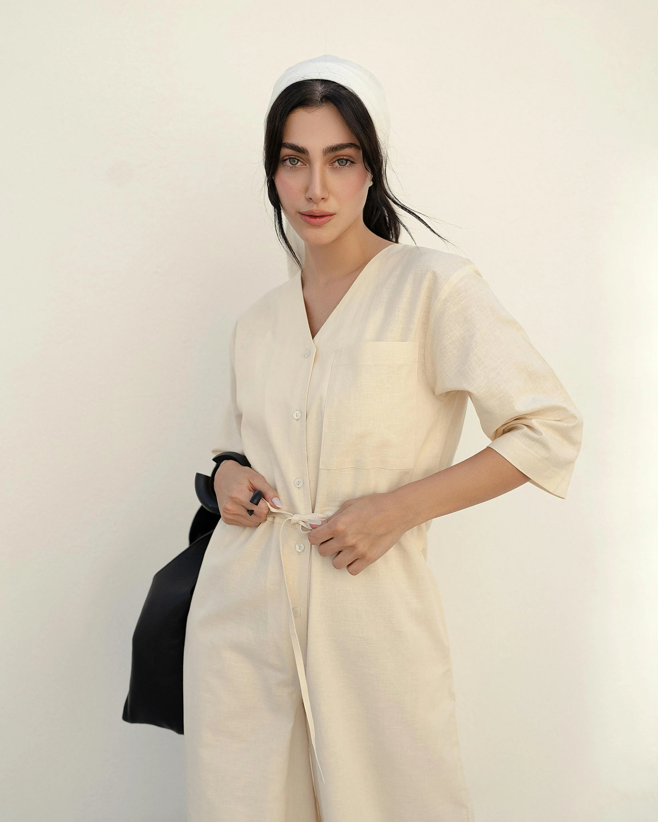 A young woman with dark hair and blue eyes wearing a cream-colored button-up dress and a white headscarf, standing against a plain off-white background.