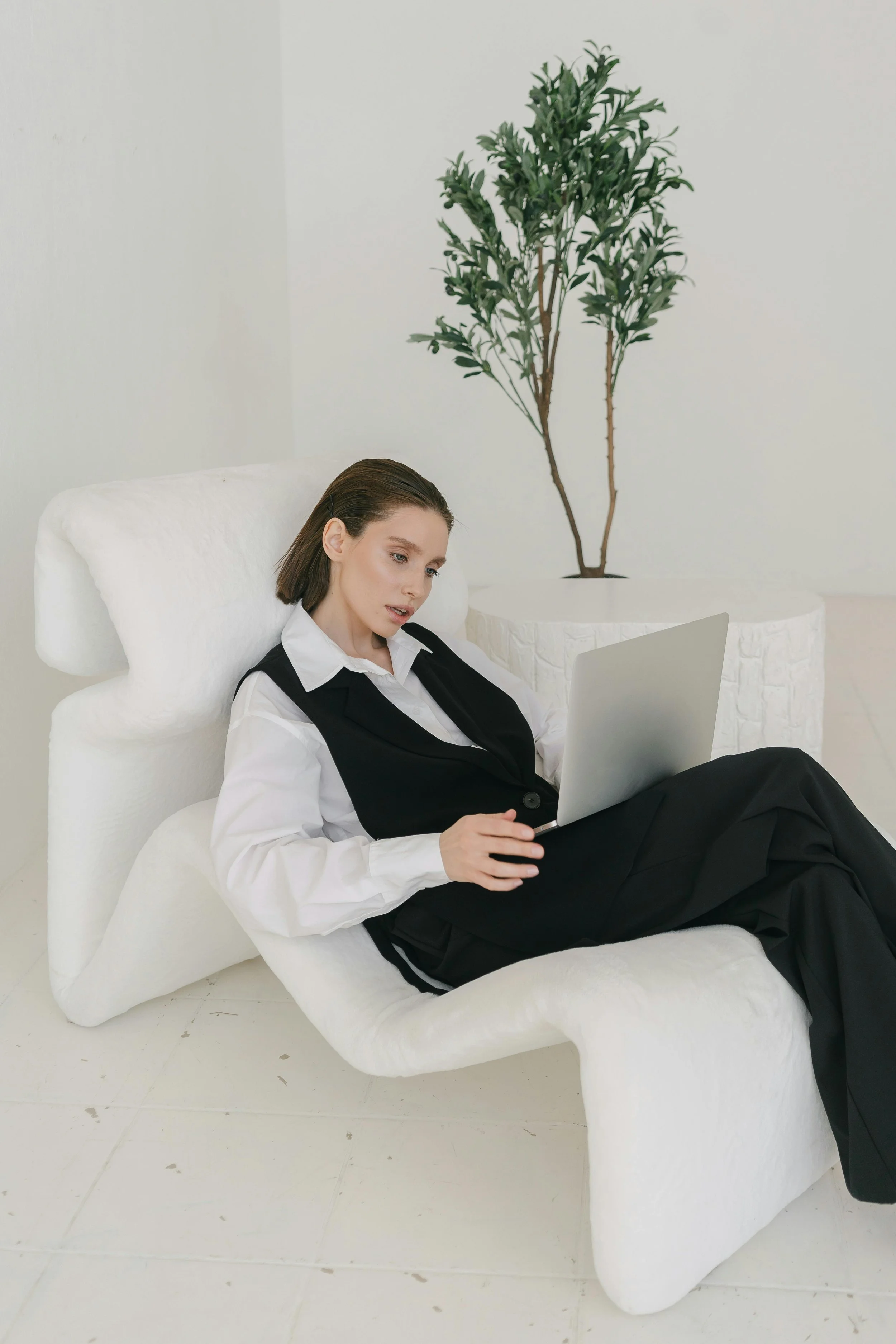 A young woman in a black vest and white shirt sitting on a modern white lounge chair, working on a silver laptop in a minimalist white room with a large green plant in the background.