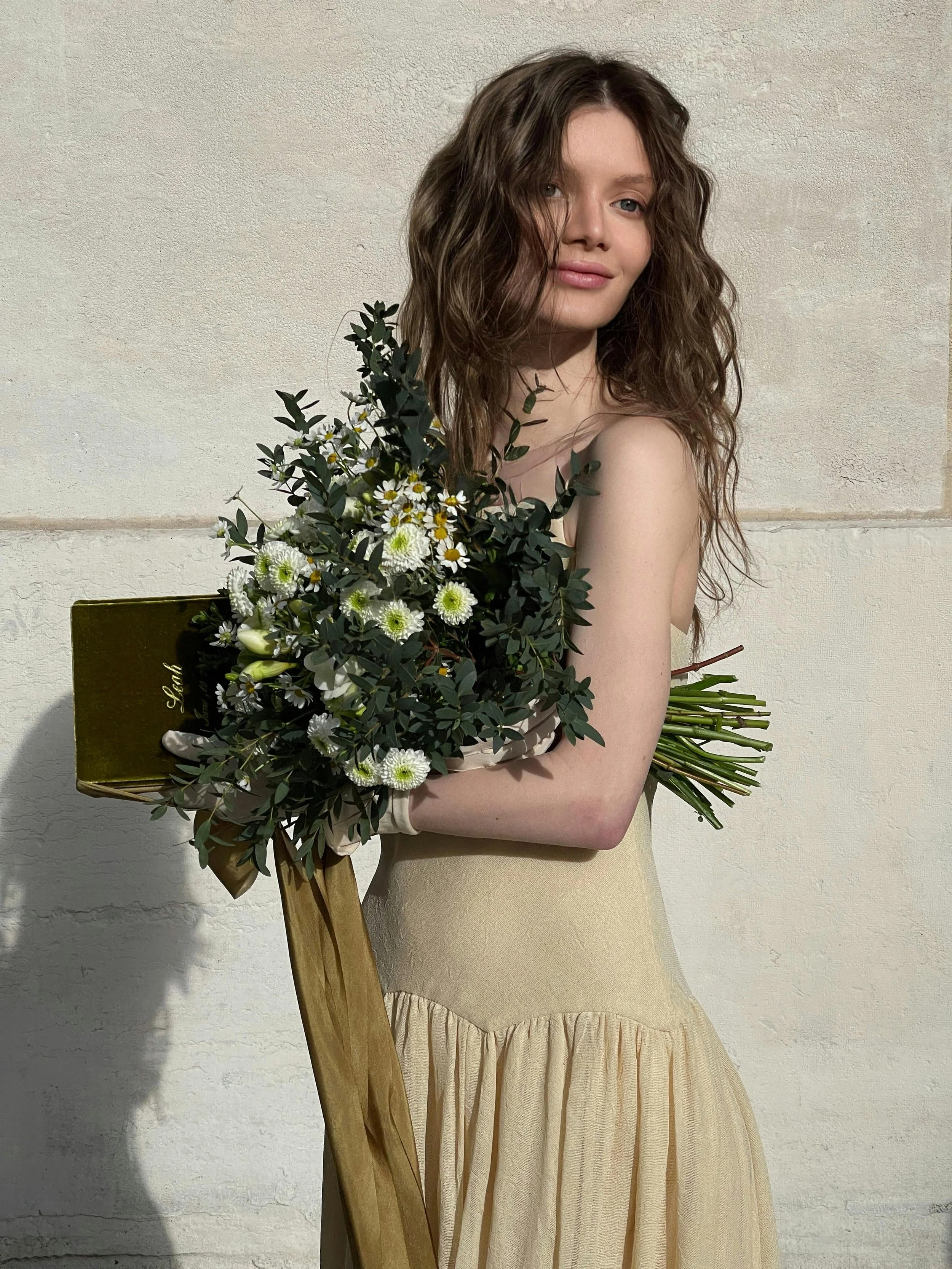A young woman with wavy, shoulder-length hair holds a bouquet of flowers and a gold clutch bag against a plain wall.
