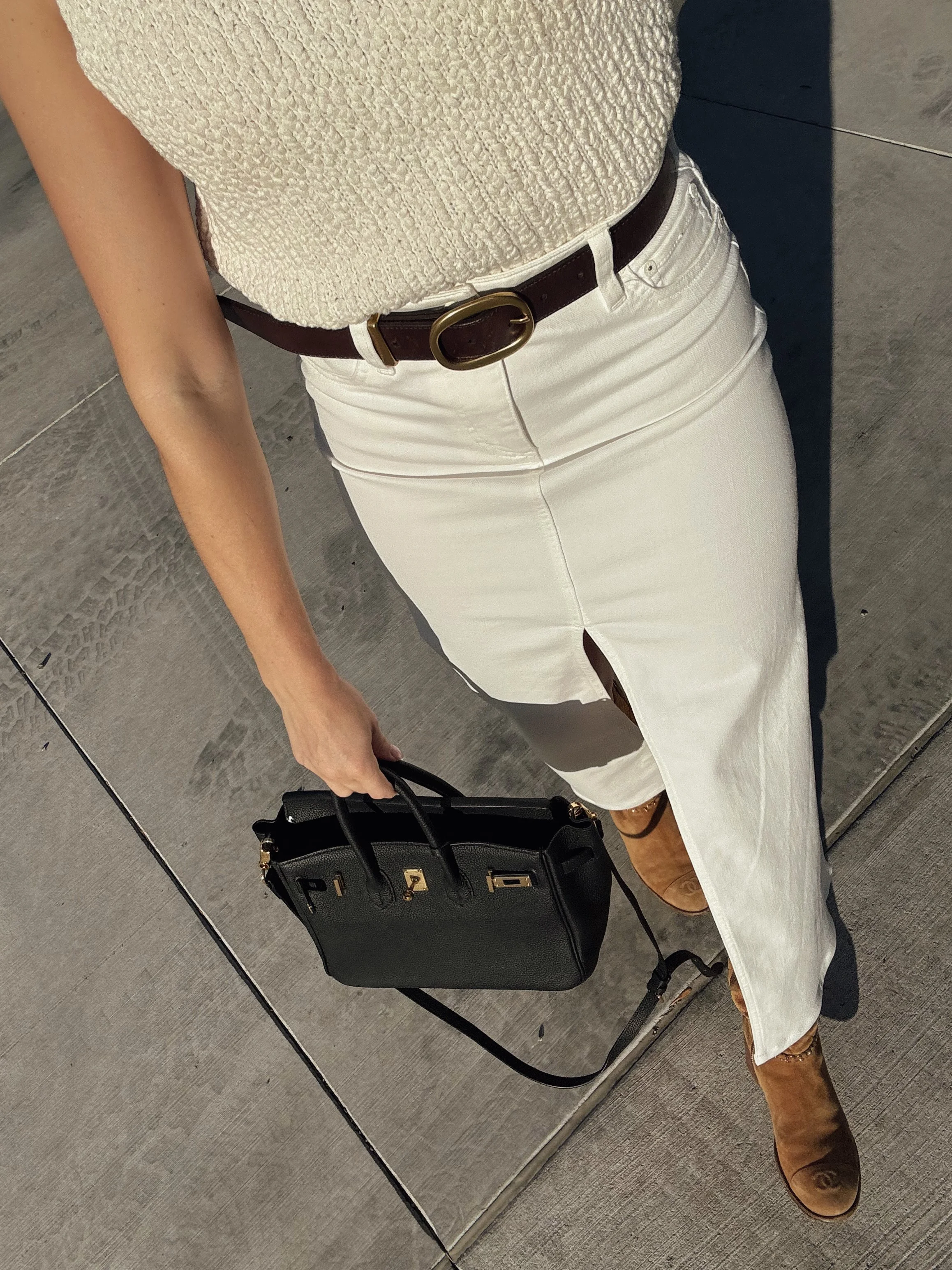 A woman dressed in a textured cream top, white skirt with a slit, brown belt, brown ankle boots, and carrying a black handbag with gold hardware, standing on a concrete sidewalk with a mirrored wall.