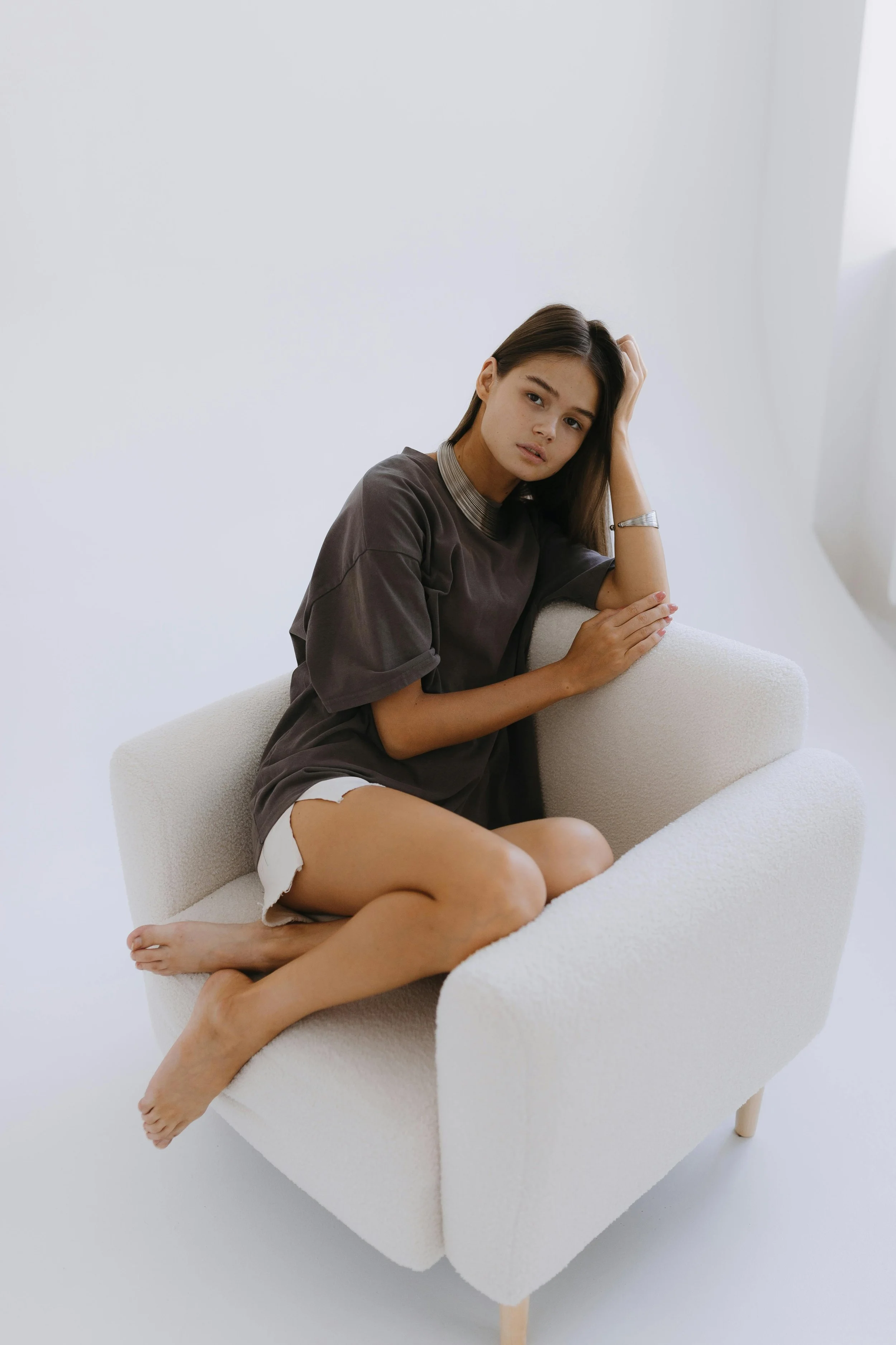 A young woman sitting on a white armchair with a neutral background, wearing a loose dark T-shirt and white shorts, with her hand resting on her head and a contemplative expression.