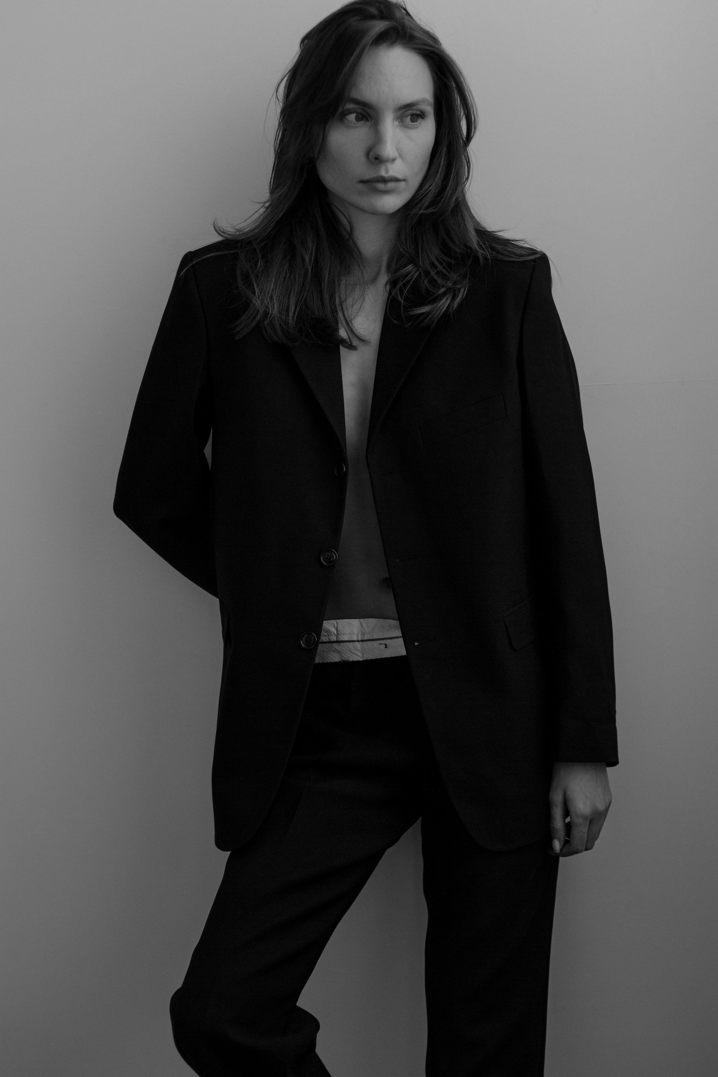 A woman with long hair wearing a black blazer and black pants stands against a plain background, gazing to her left.