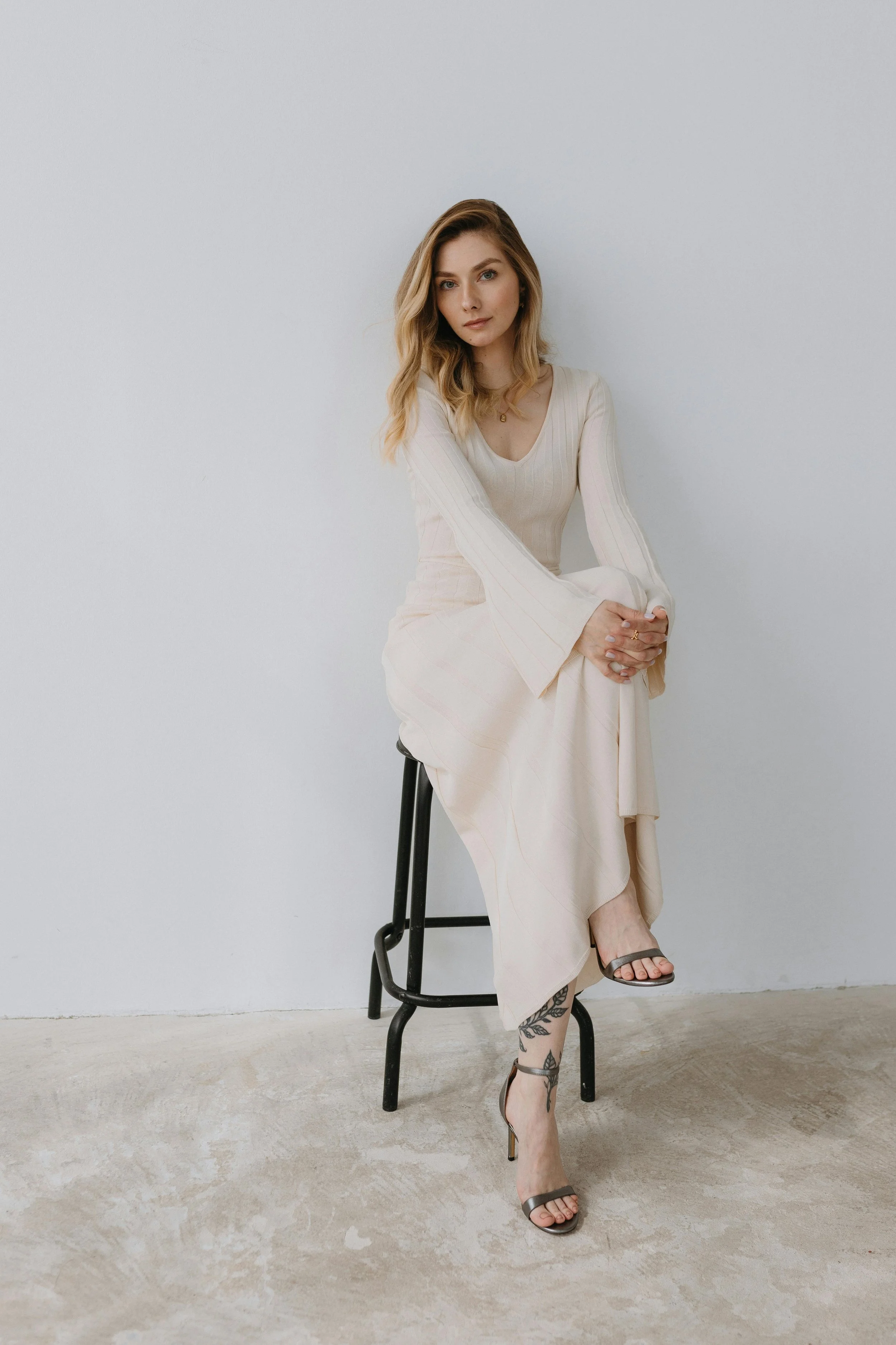 A woman with shoulder-length blonde hair sitting on a black stool against a plain white wall. She is wearing a long, beige dress with bell sleeves and black high-heeled sandals. She has a tattoo on her left ankle and is looking directly at the camera with a neutral expression.